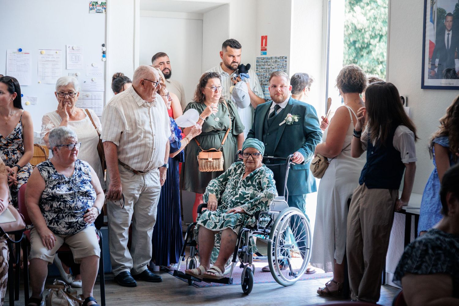 Wedding guests gathered in a hall, including an elderly woman in a wheelchair, dressed in formal attire for a celebration.