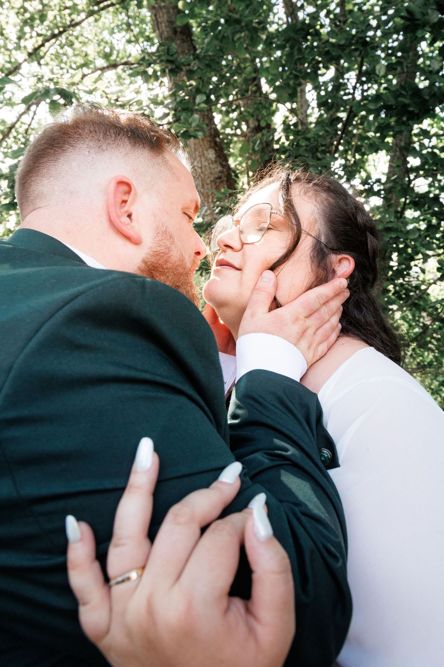 Groom tenderly cups bride's face for a kiss under lush green trees at outdoor wedding.