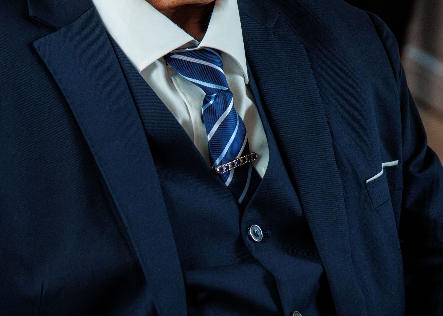 Man in a navy blue three-piece suit with a striped blue and white tie secured by a gold tie bar.