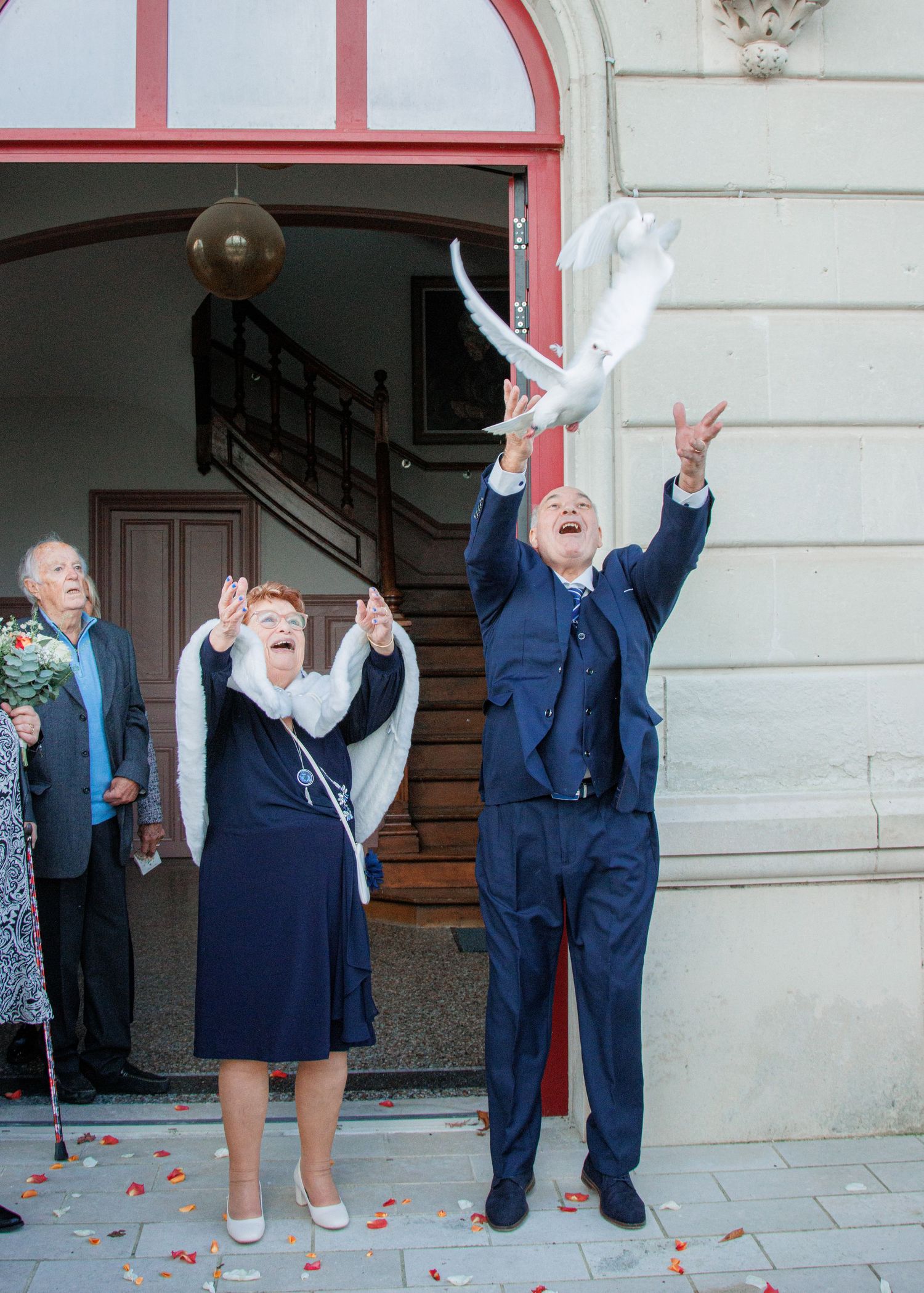 Elderly couple releasing white doves outside a building entrance during a wedding celebration.