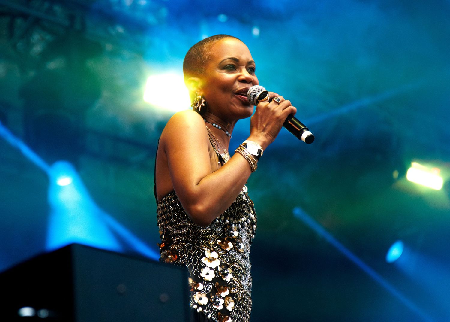 A performer sings passionately on stage wearing a sparkly silver dress against a blue-lit backdrop during a concert performance.