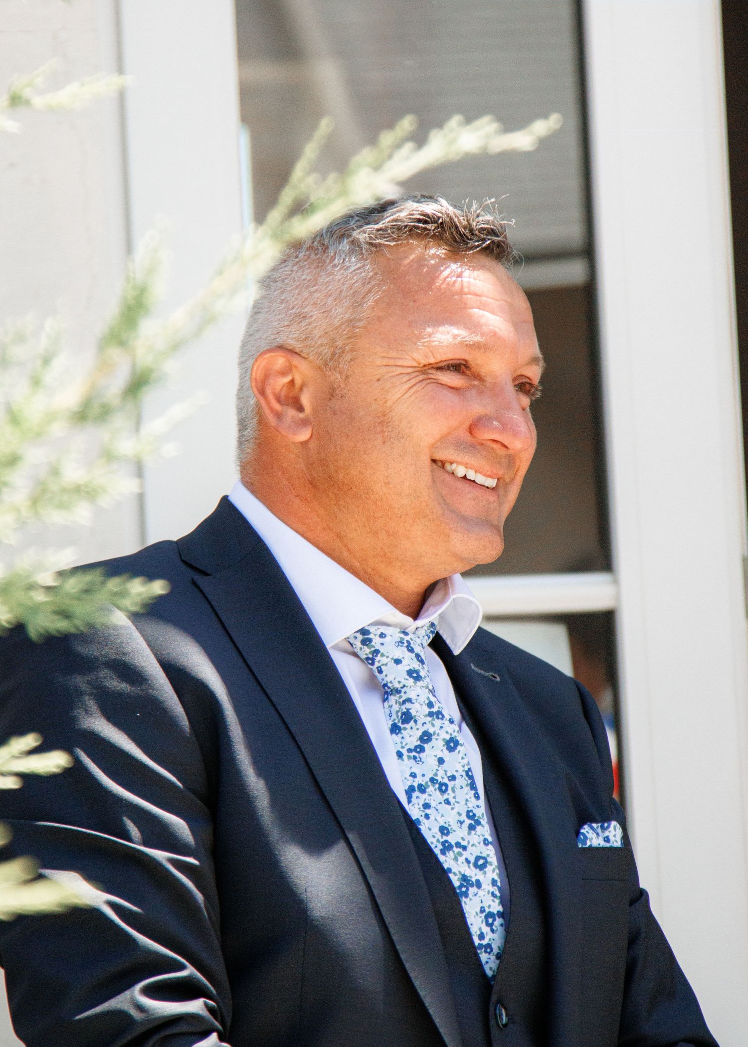A person in a dark suit and blue floral tie smiling outdoors on a sunny day.