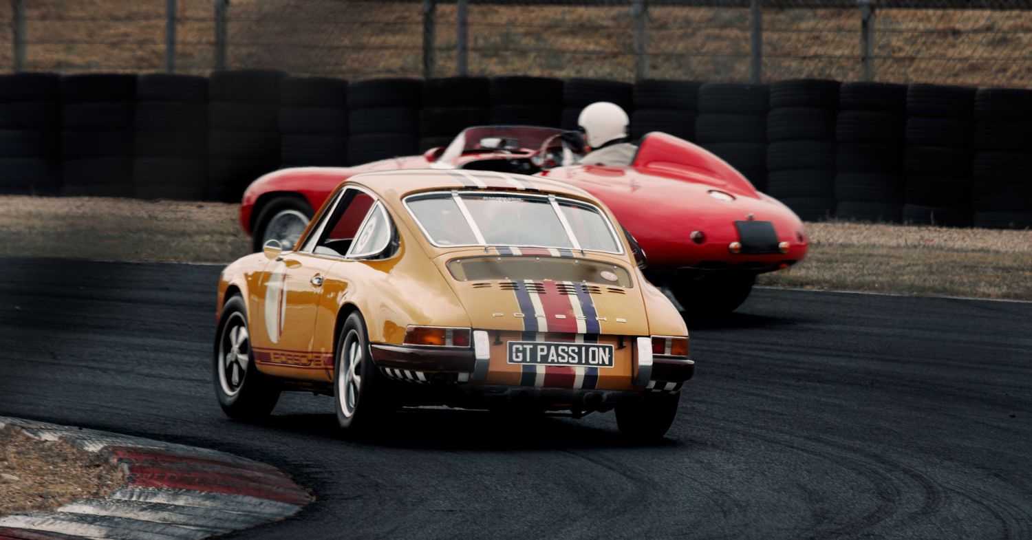 Vintage sports cars including a red racer compete on a professional racing circuit.