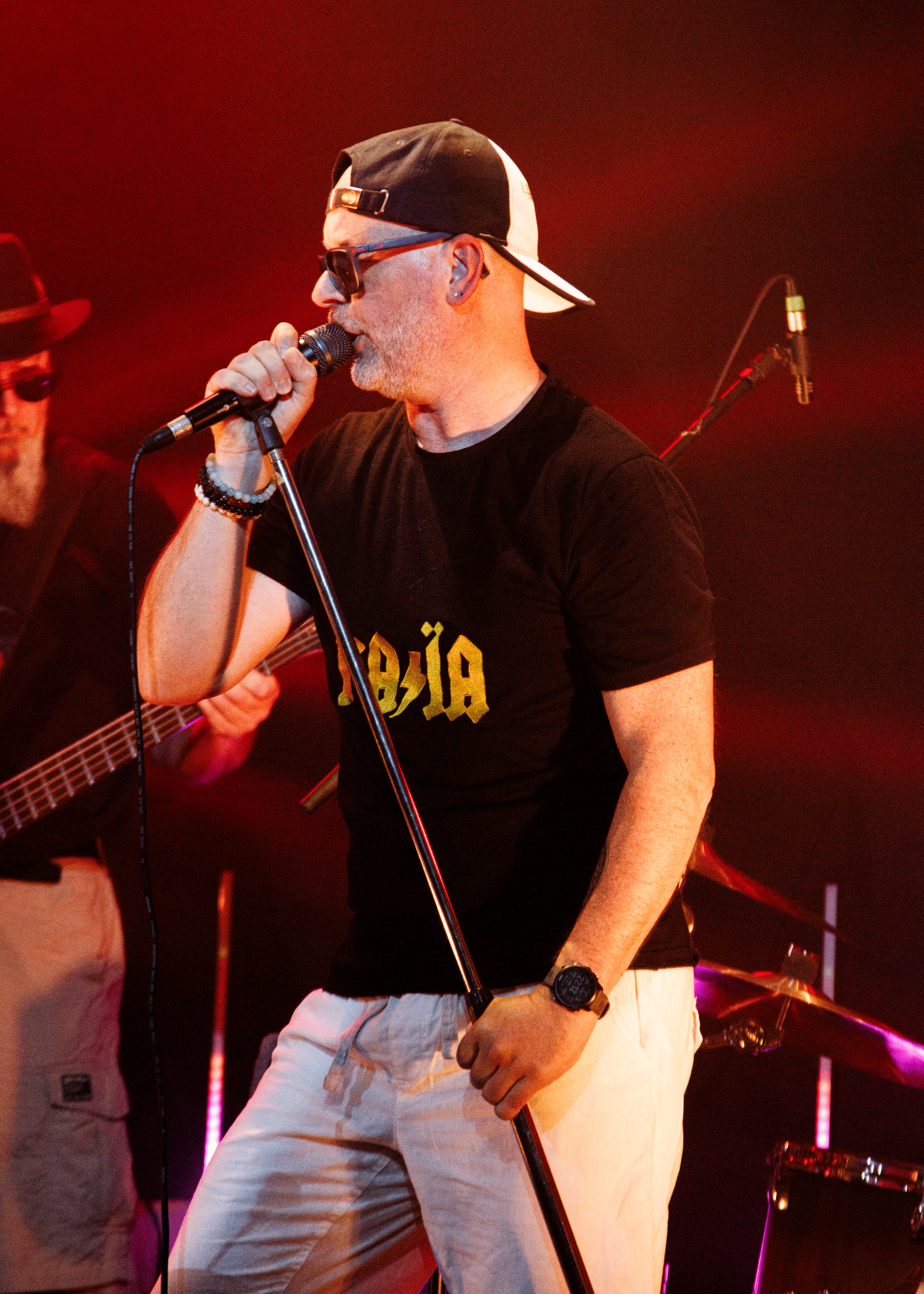A vocalist performs energetically on stage wearing a black t-shirt and baseball cap under dramatic red lighting.