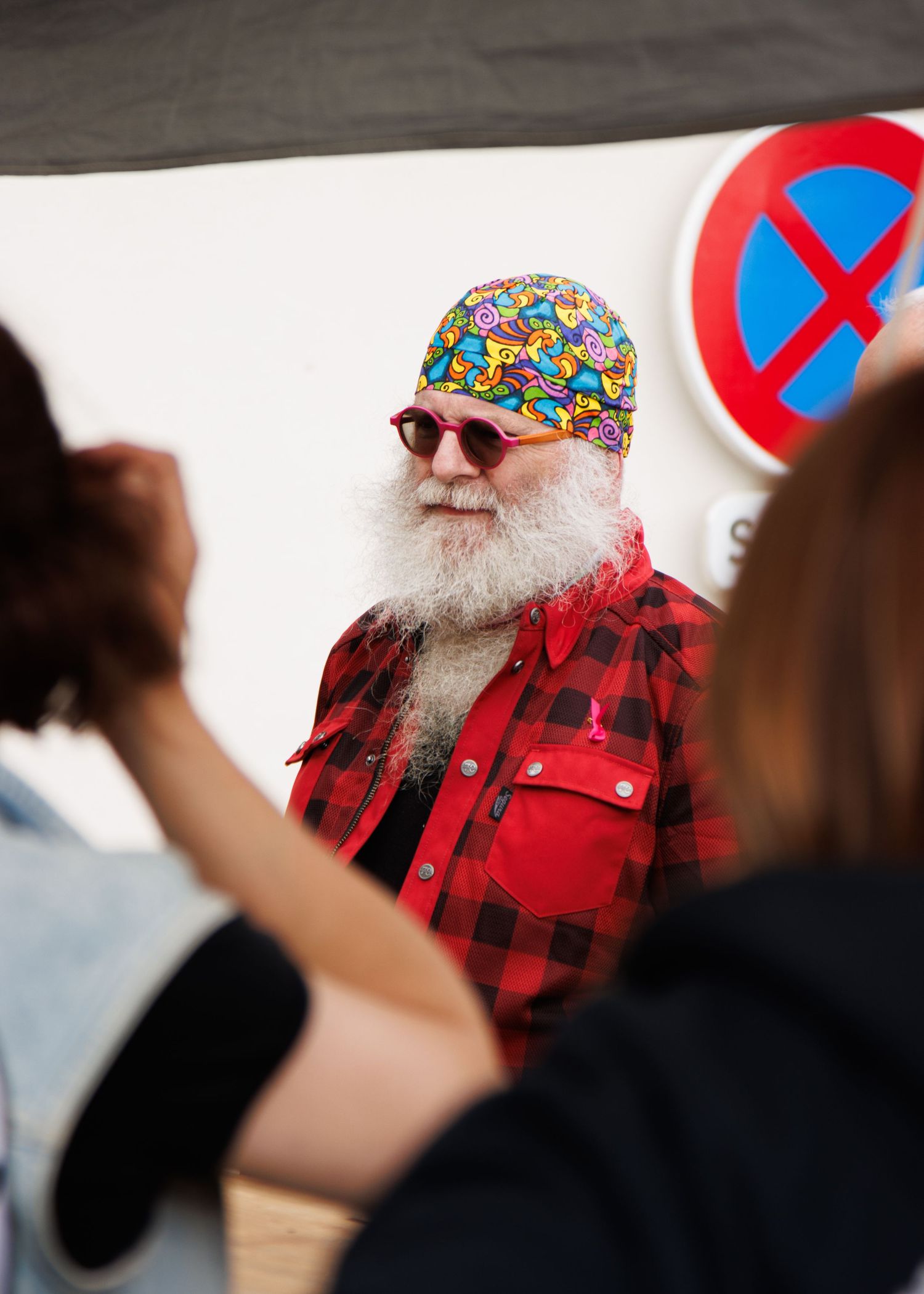 Person with white beard wearing red plaid shirt and colorful bandana poses for photos with crowd.