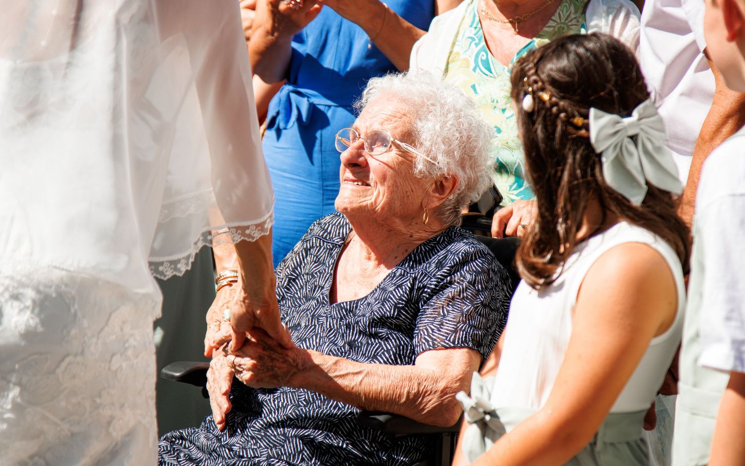 Elderly woman in a navy blue dress smiles while sitting among a crowd at a celebratory event.