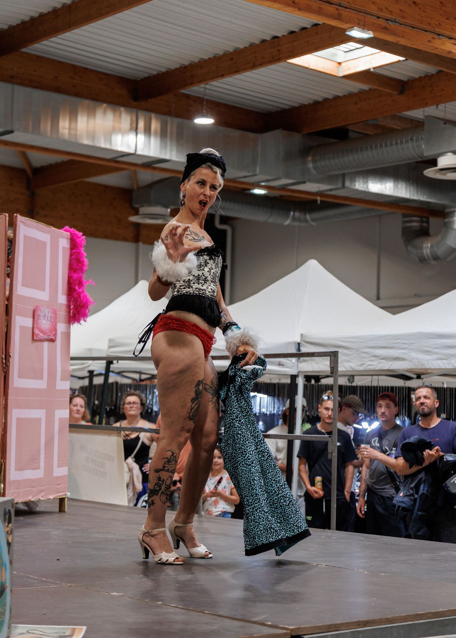 A tattooed burlesque performer in lingerie and heels struts on stage at an indoor event, holding a garment before an audience.