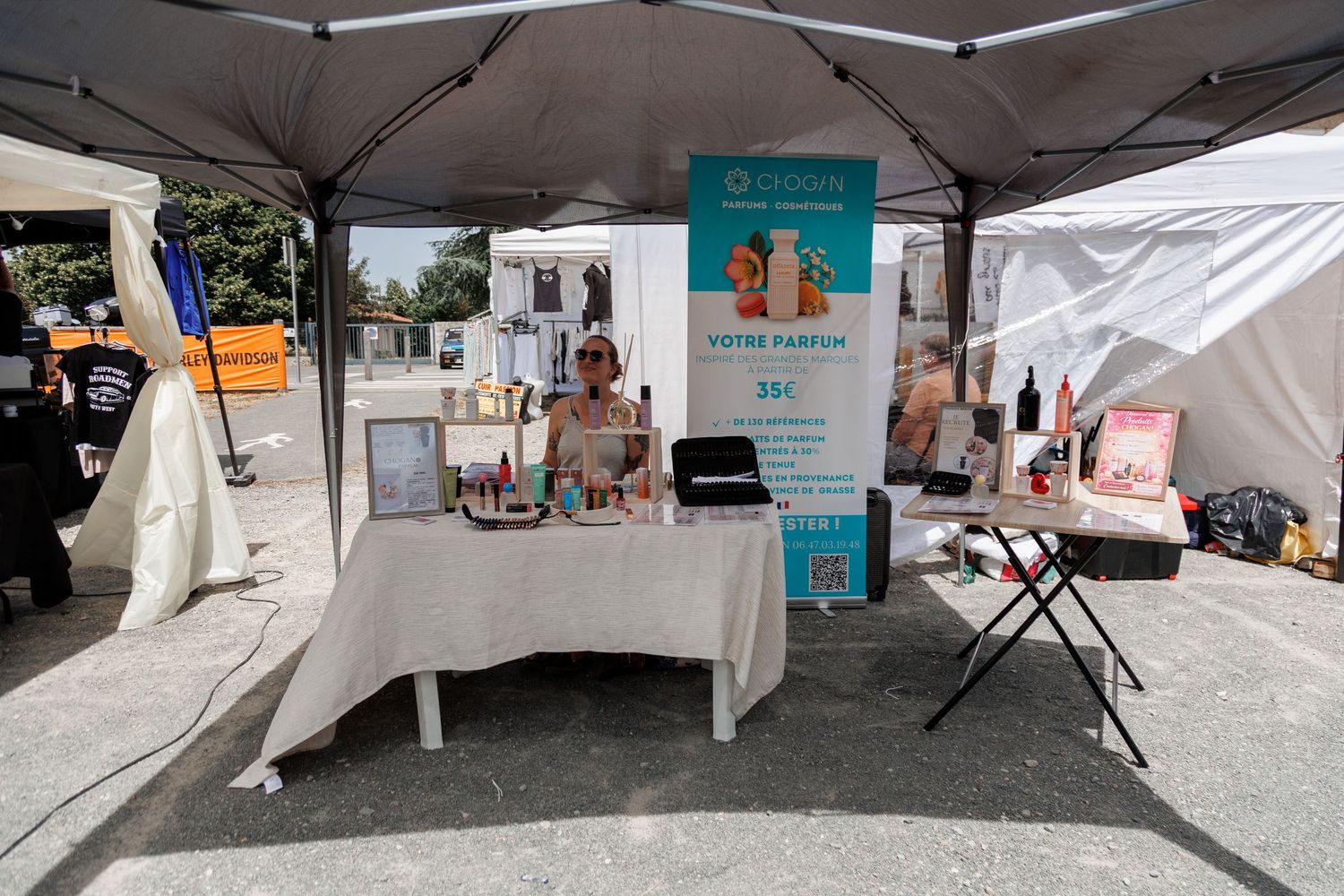 Outdoor market booth under gray canopy selling custom perfumes with white banner and product display table.