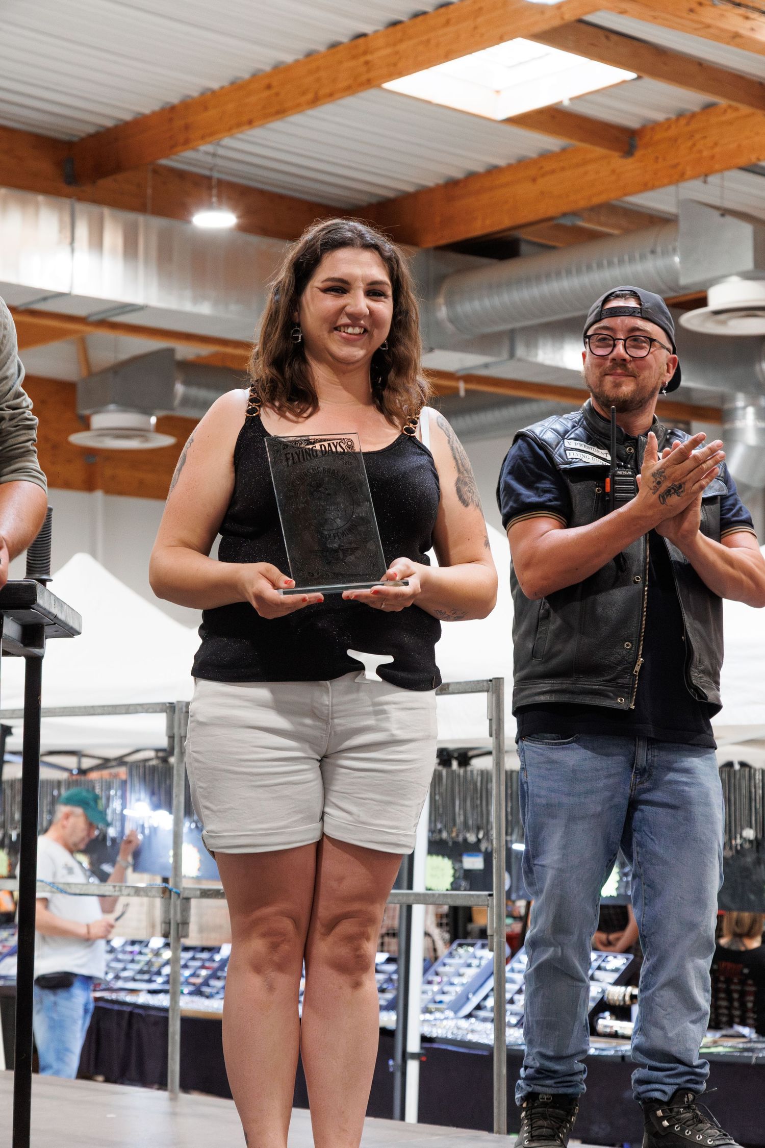 Woman proudly holds award plaque while man in leather jacket applauds at indoor ceremony.