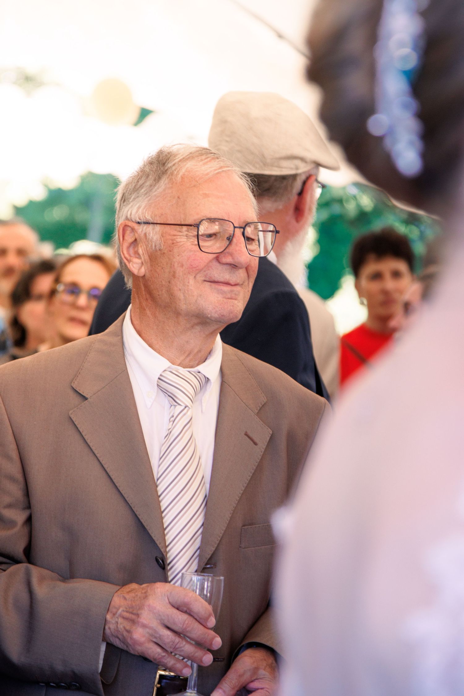 Senior man in beige suit and striped tie holding a glass at a wedding reception gathering.