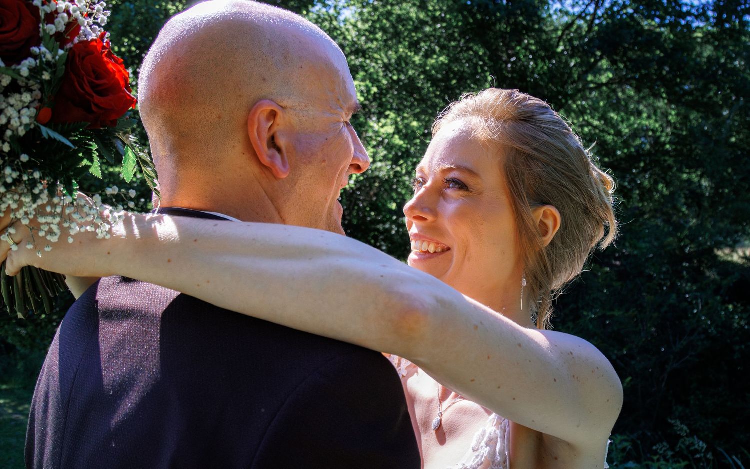 Bride and bald groom share a romantic embrace outdoors surrounded by lush green trees and red roses.