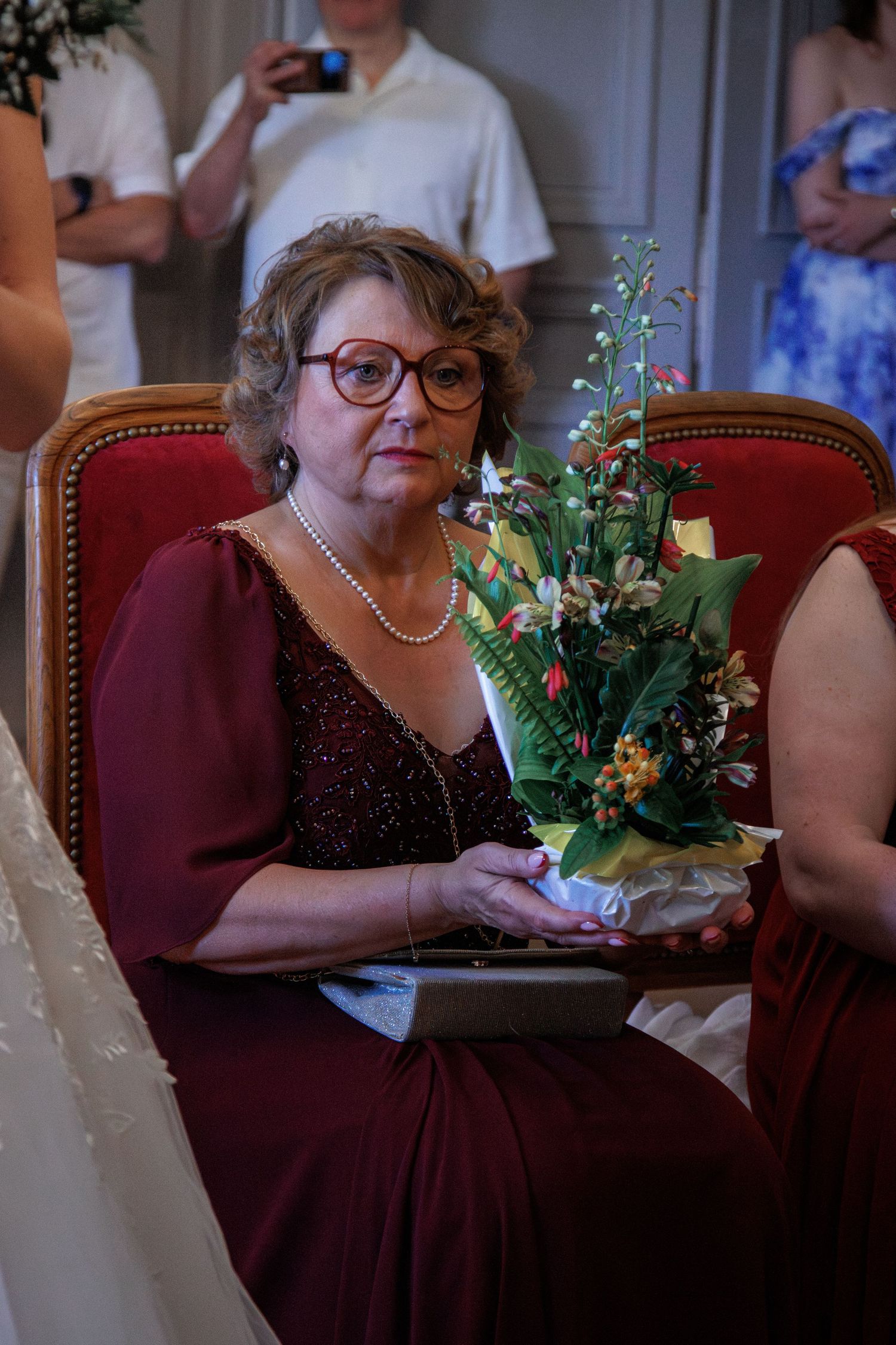 Elegant older woman in burgundy dress with pearl necklace holding a floral bouquet at a wedding.