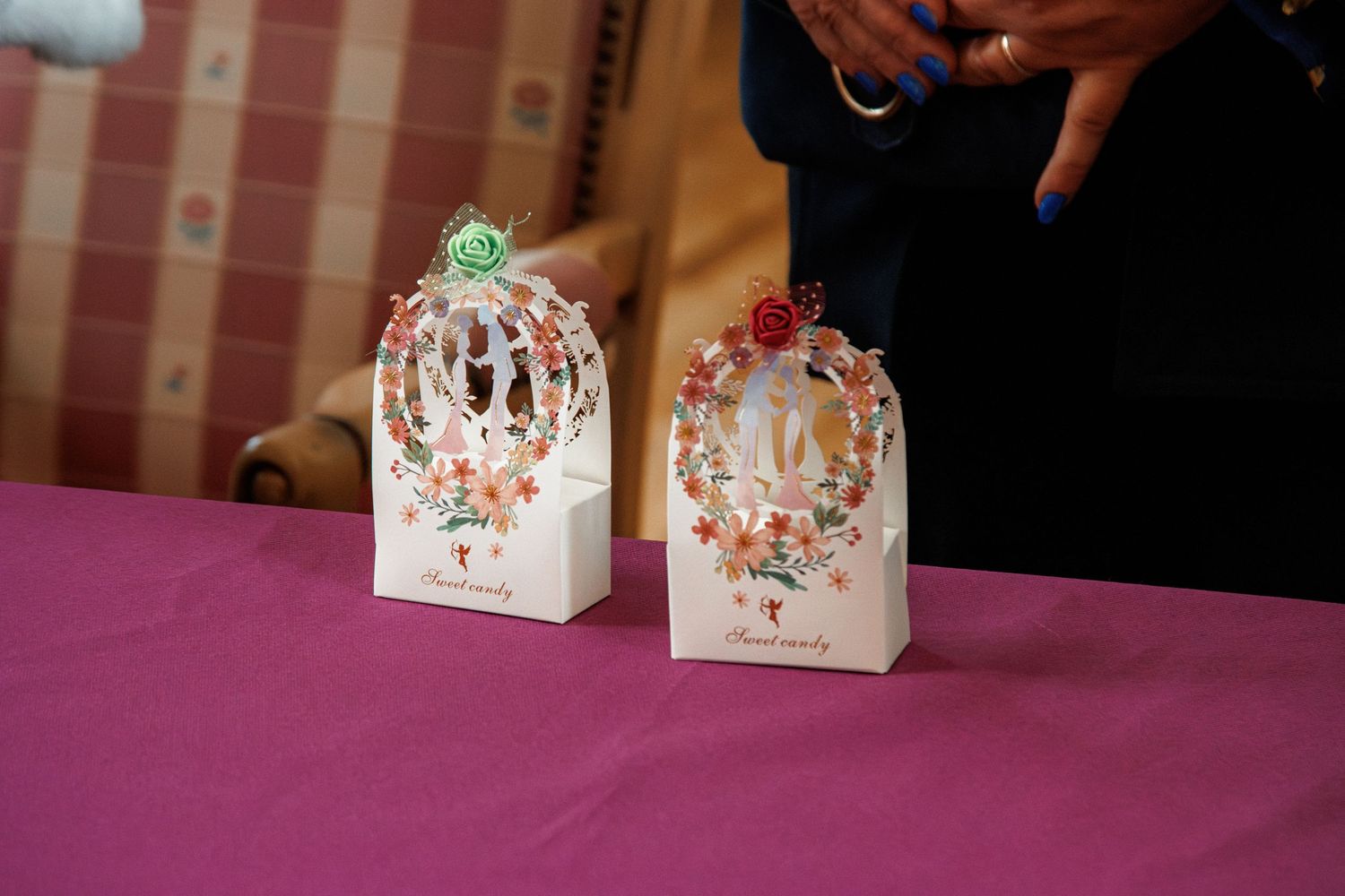 Two decorative white gift boxes with floral cutout designs and rose toppers displayed on a pink tablecloth.