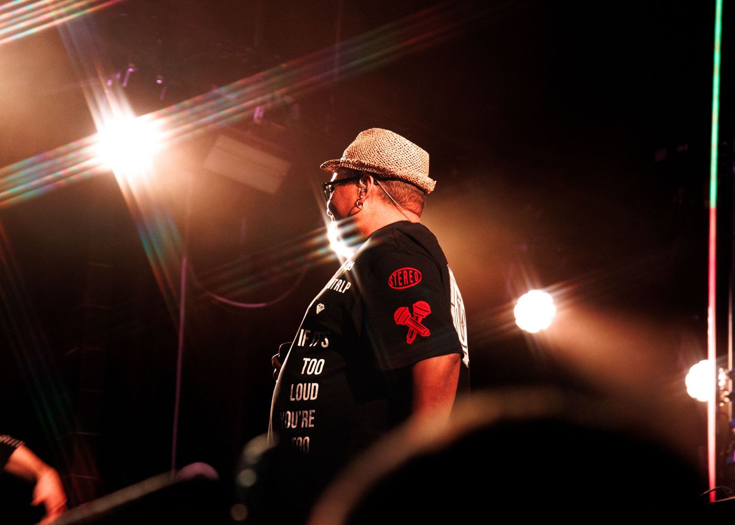 A performer wearing sunglasses and a white hat stands on stage with dramatic lighting and lens flares in the background.
