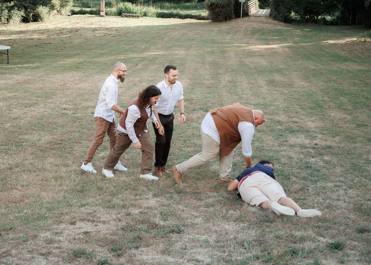 Group of wedding guests playfully interact on a lawn, with one person lying on the grass during outdoor celebration.