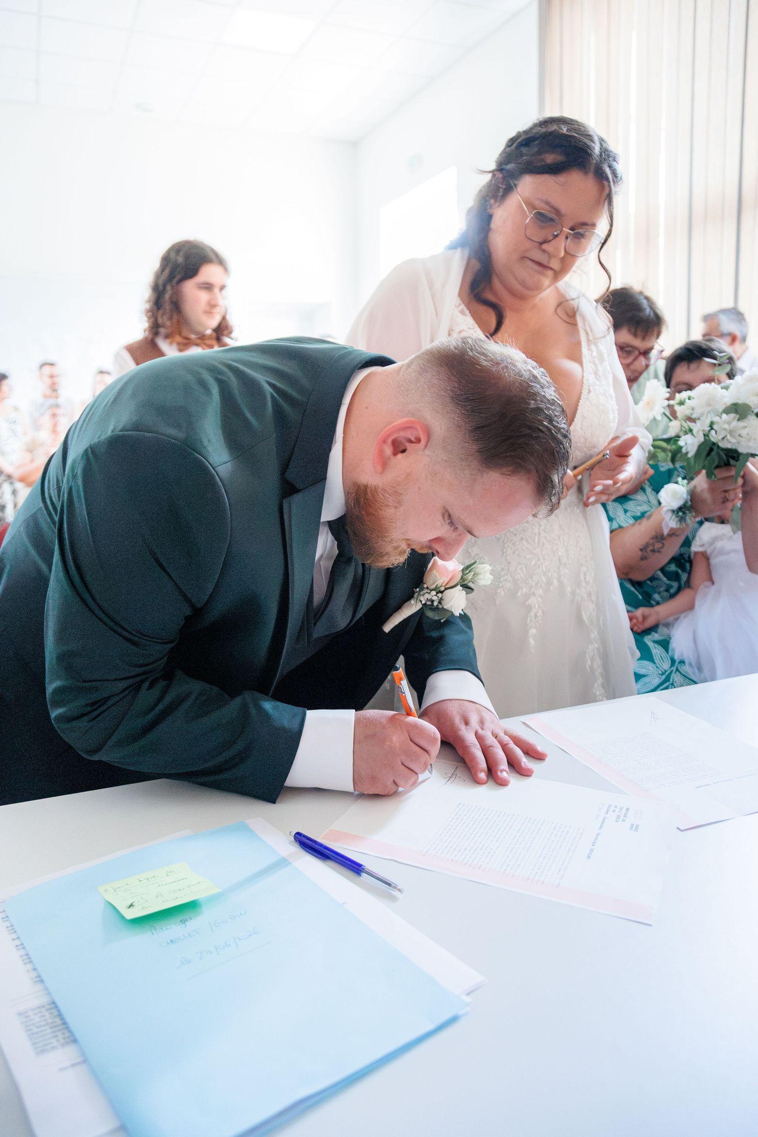 Groom in dark suit signs wedding documents at civil ceremony while bride watches.