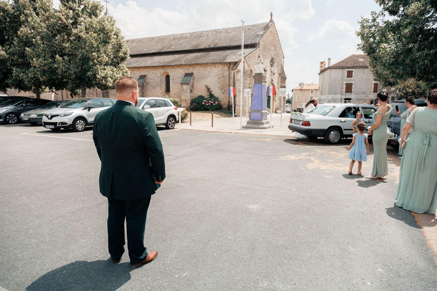 People standing in a sunny town square near a church with parked cars on a warm summer day.