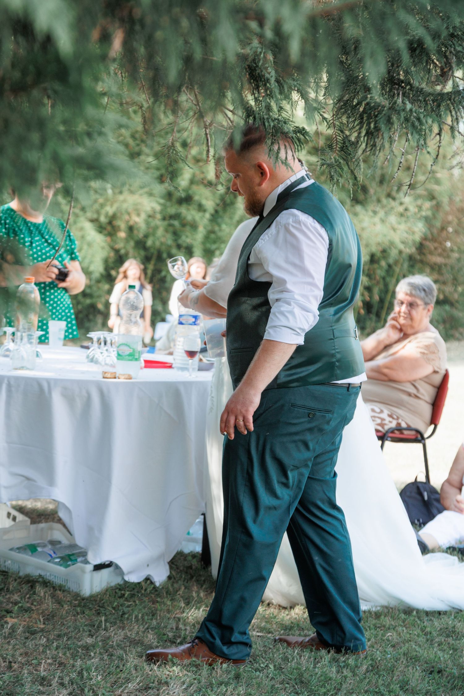 Bearded groom in green vest and trousers stands near outdoor wedding reception table under pine trees.