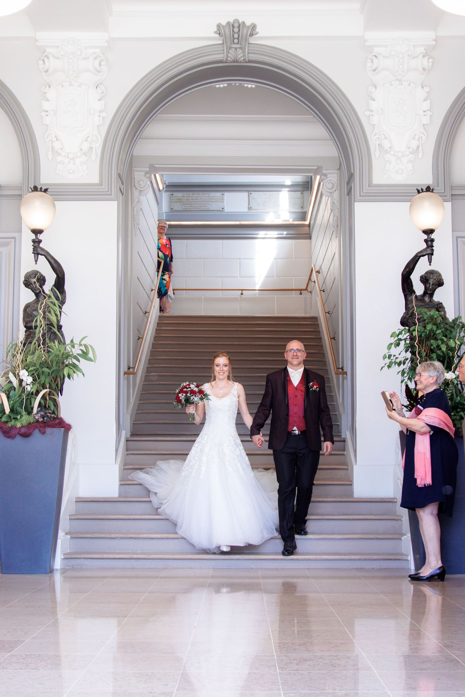 Newlyweds descend a grand staircase hand in hand as a guest photographs them at their wedding.