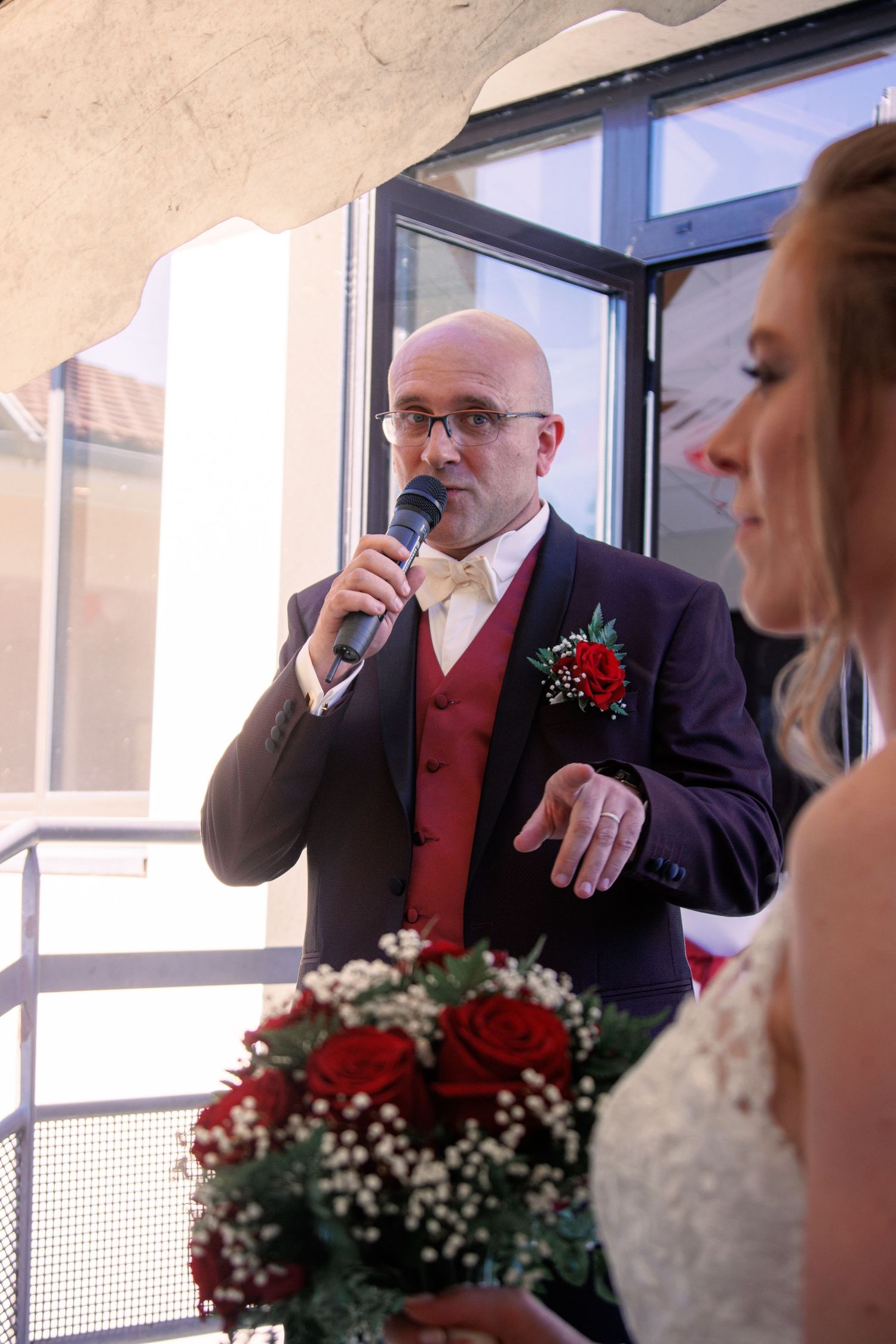 Bald groom in purple suit speaks into microphone at wedding ceremony, bride holding red rose bouquet in foreground.