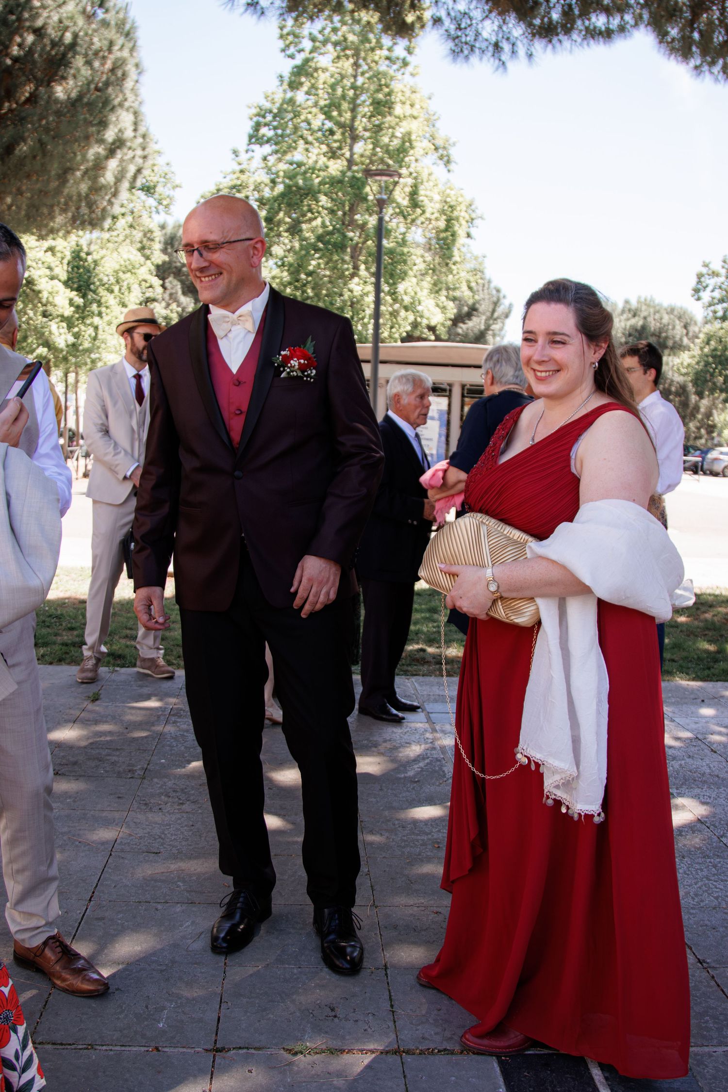Elegantly dressed wedding guests in formal attire walk outdoors, woman in red gown with white shawl.