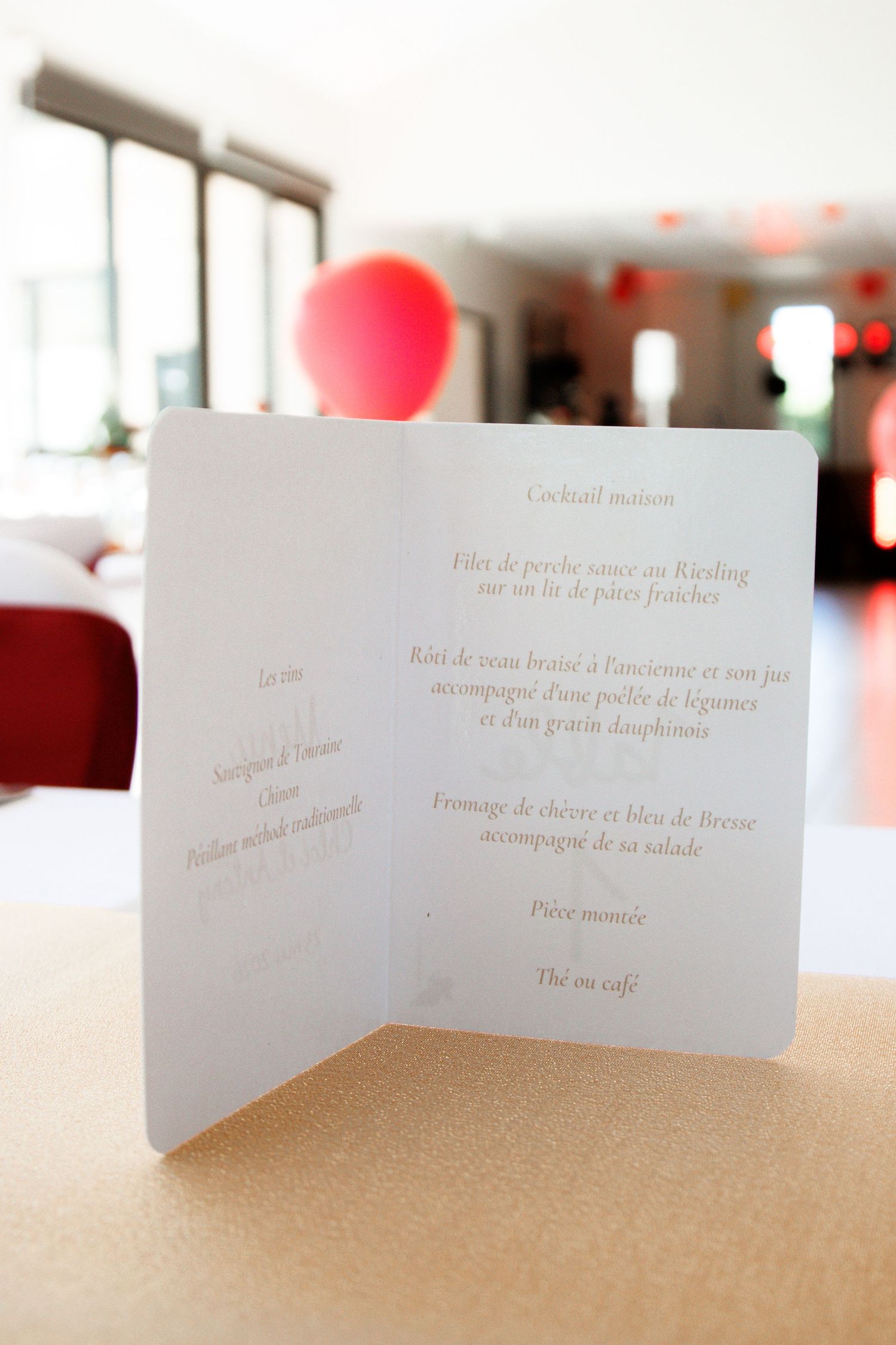 Elegant French wedding menu card standing on a table with red balloons visible in the blurred background.
