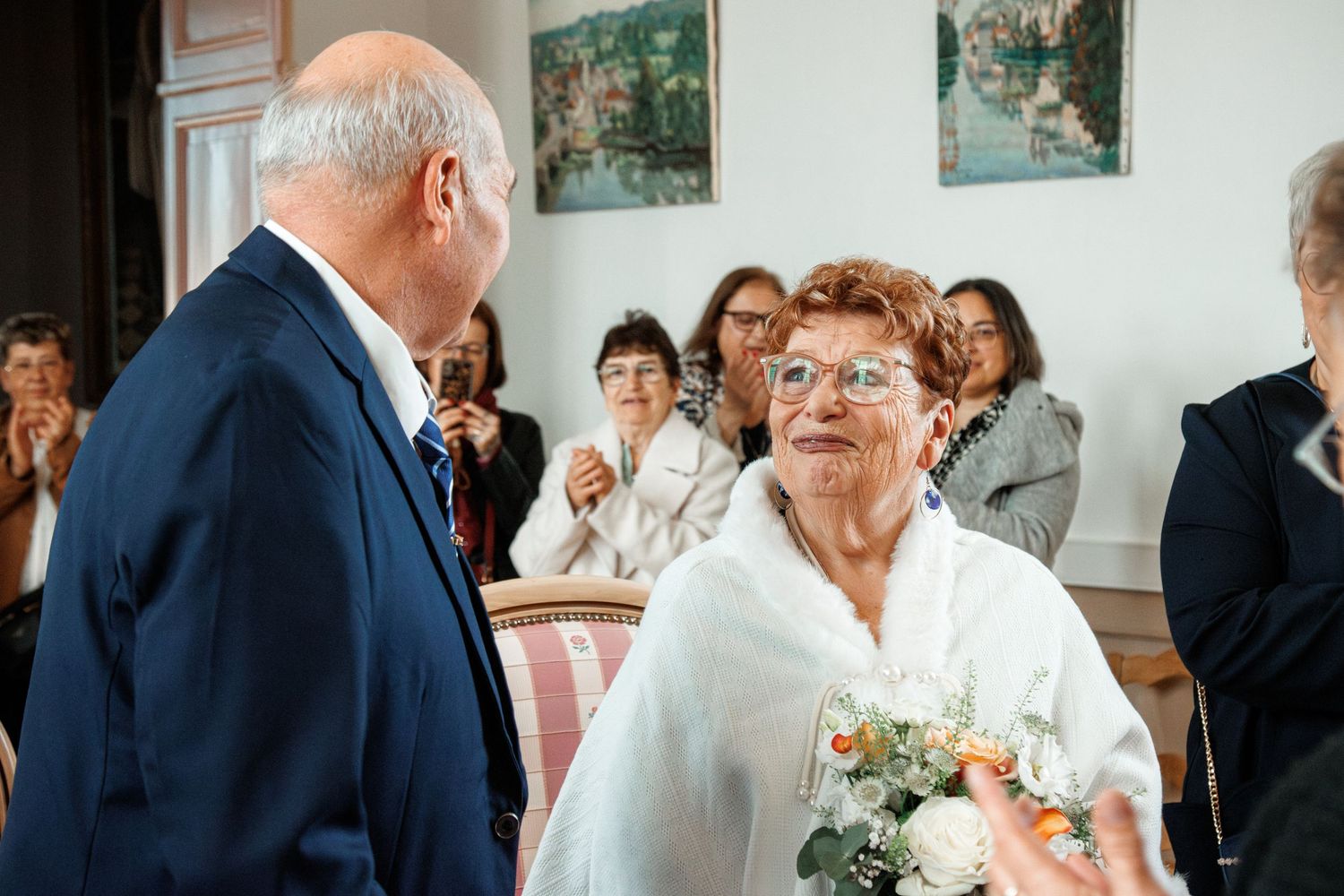 Elderly couple sharing a tender moment at a wedding ceremony, woman holding a bouquet, guests applauding in background.