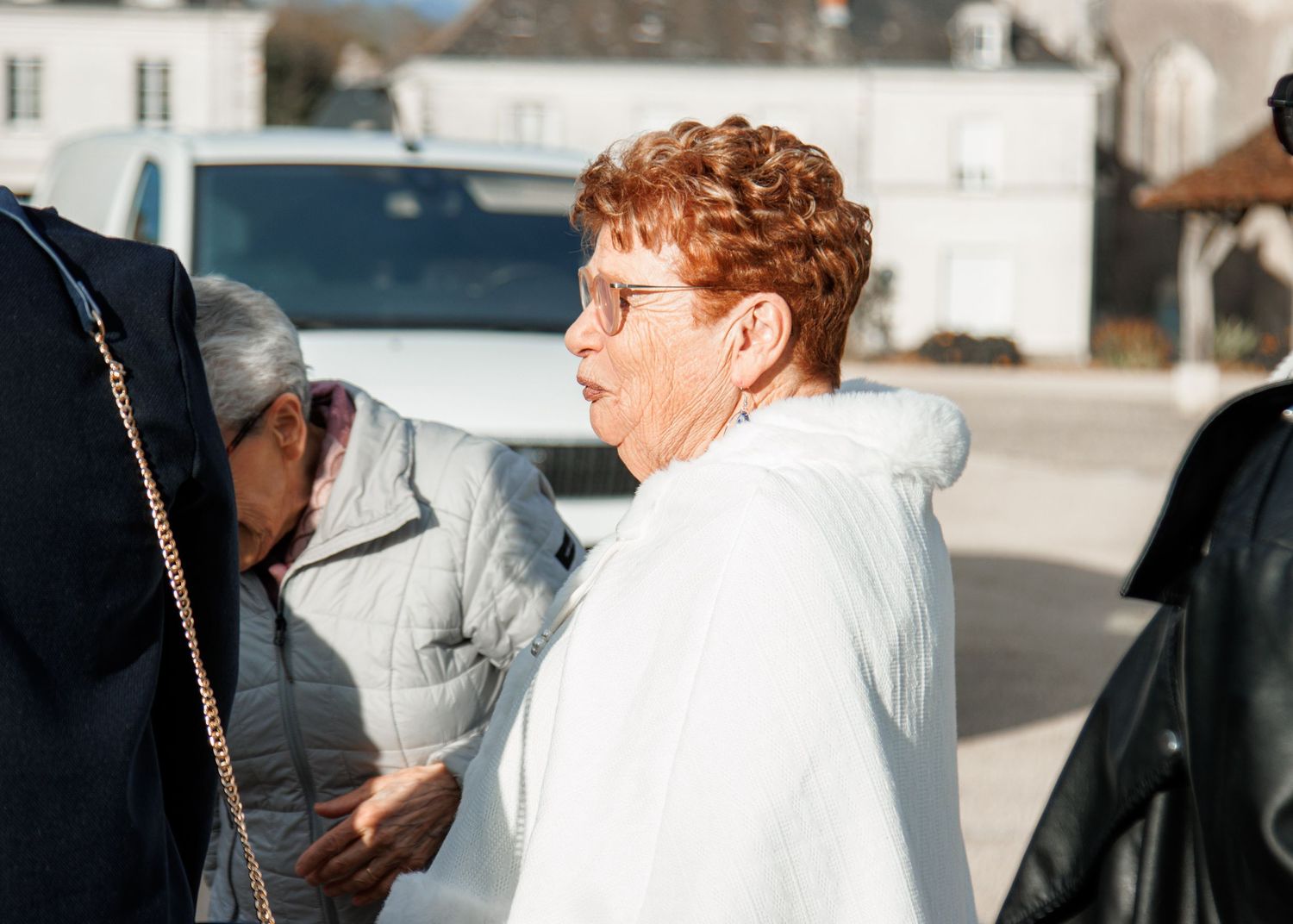 Two elderly women conversing outdoors on a sunny day, one wearing a white jacket and red curly hair.