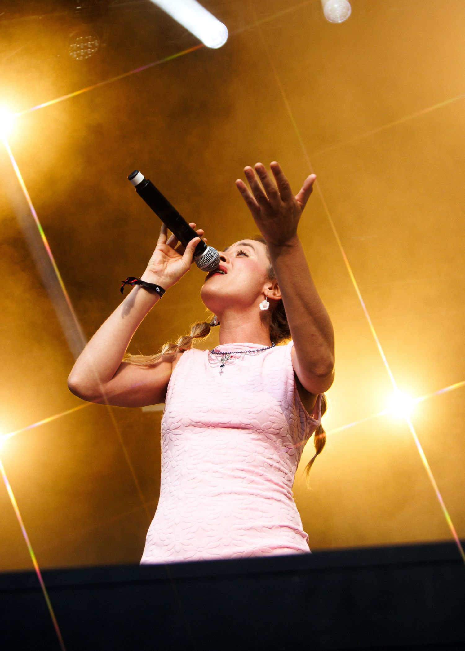 A performer in a pink tank top sings passionately on stage with dramatic golden lighting and spotlights in the background.