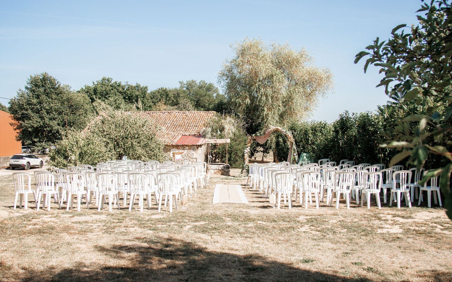 Rustic outdoor wedding ceremony setup with white chairs and a natural arch in a sunny garden setting.