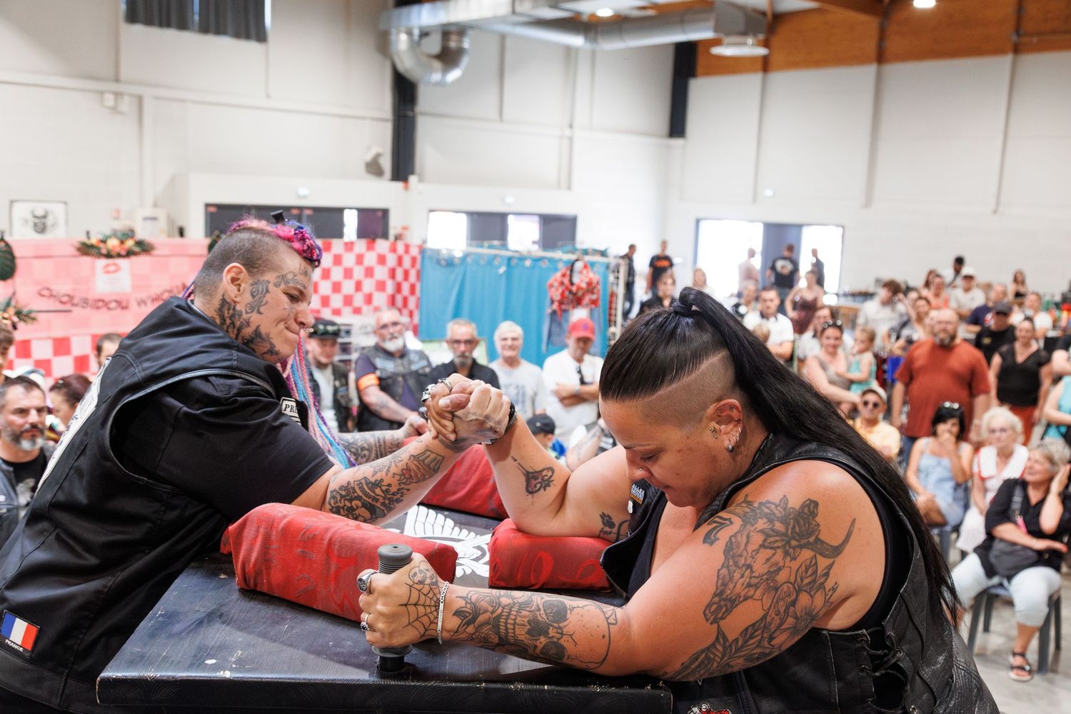 Two heavily tattooed men compete in an arm wrestling match at a crowded indoor tournament event.