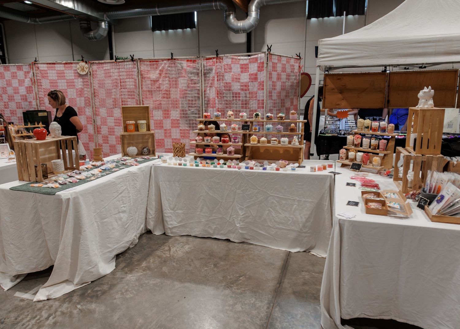 Vendor booth with white tablecloths displaying handmade goods, jars, and crafts against a pink checkered backdrop.