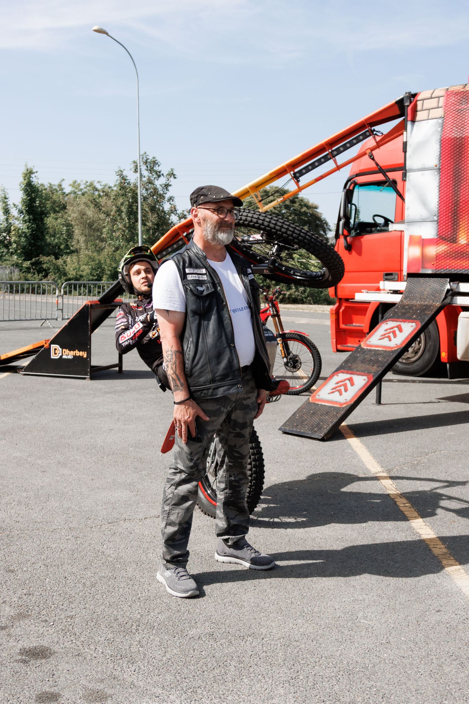 A tattooed biker carries a dirt bike on his shoulder near a red truck and ramps at an outdoor stunt event.