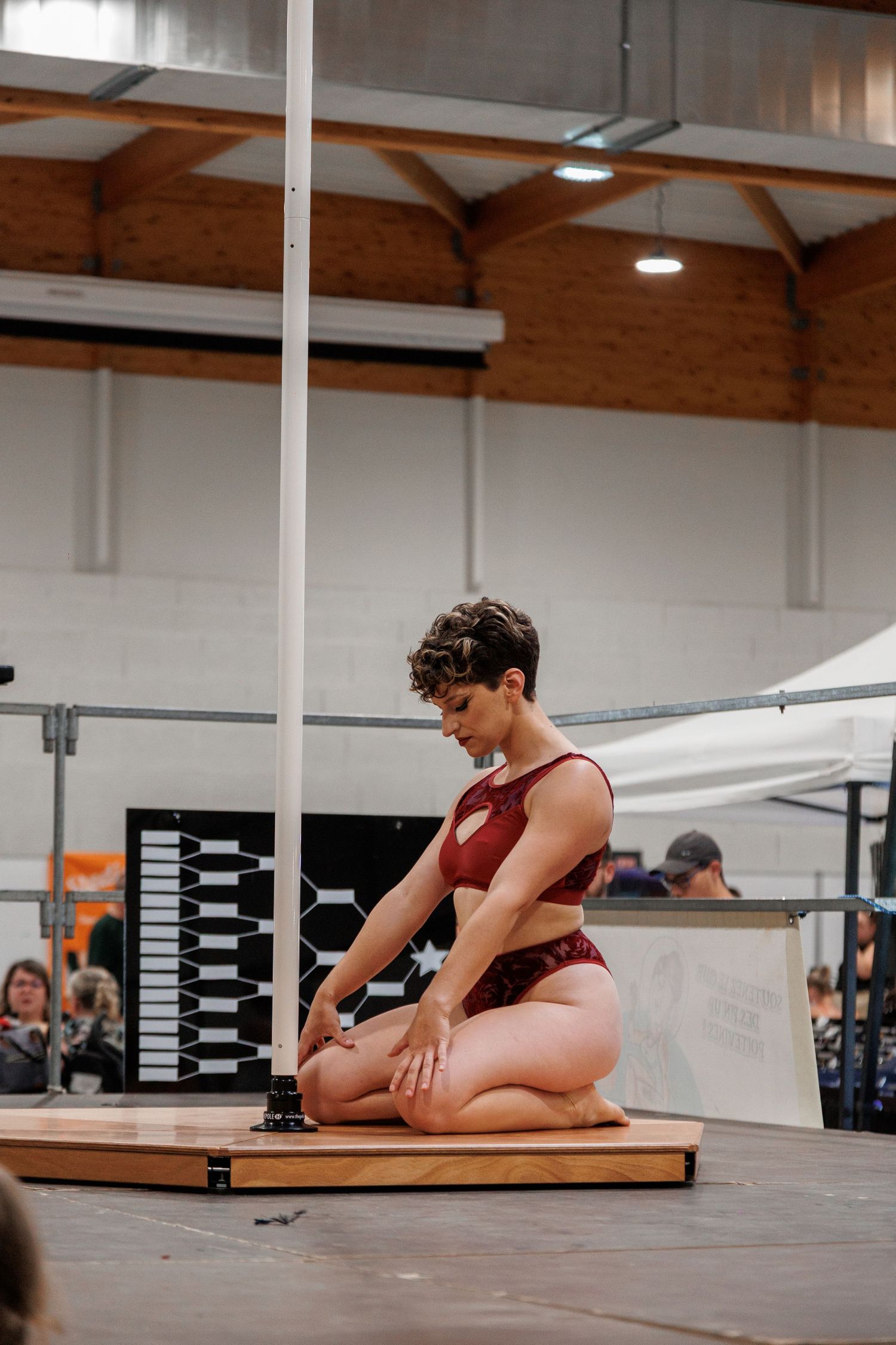 A female pole dancer in a red outfit kneels gracefully on a wooden stage platform during an indoor competition.