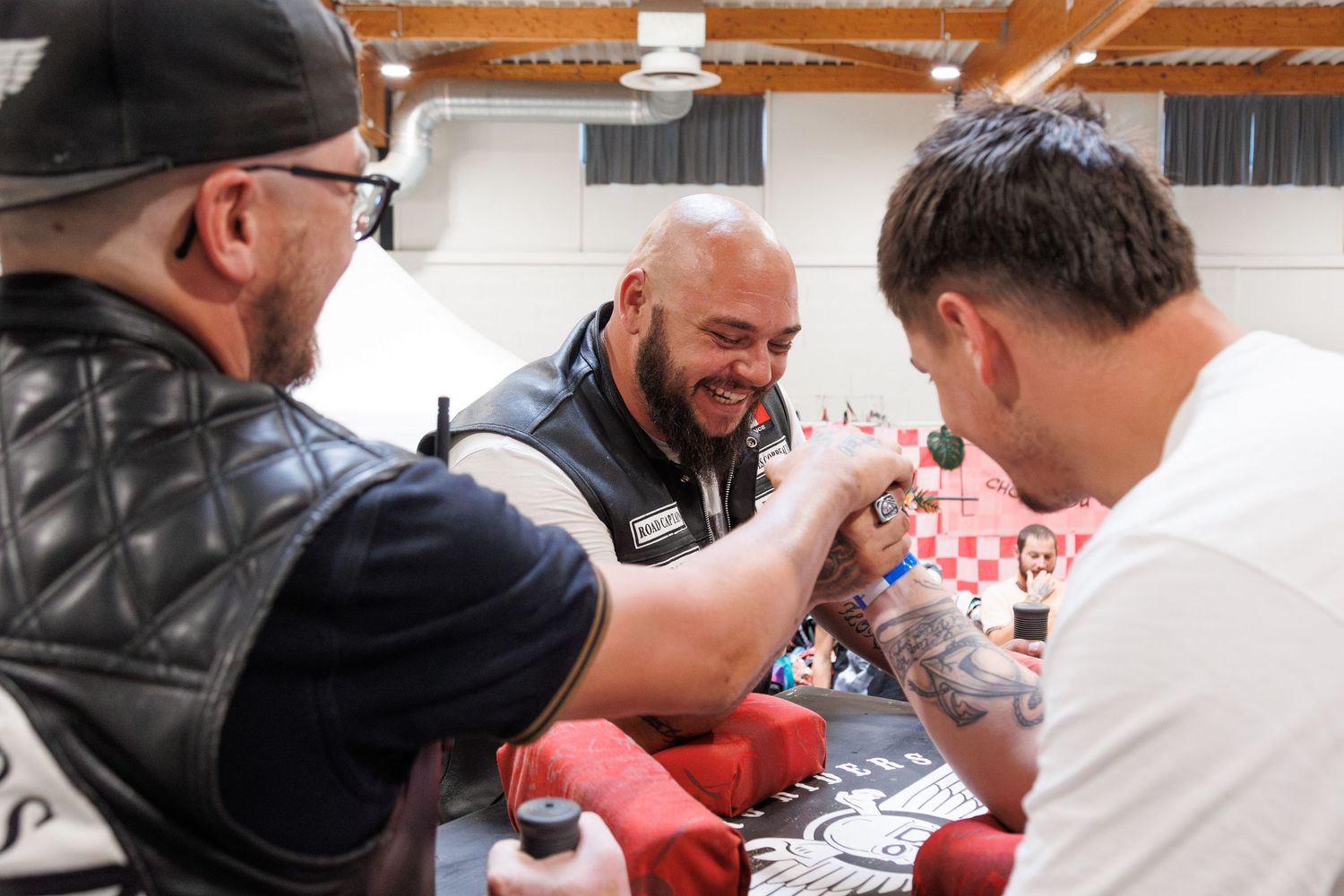 Three men arm wrestling at an indoor event, smiling and laughing, one wearing a leather vest with tattoos visible.