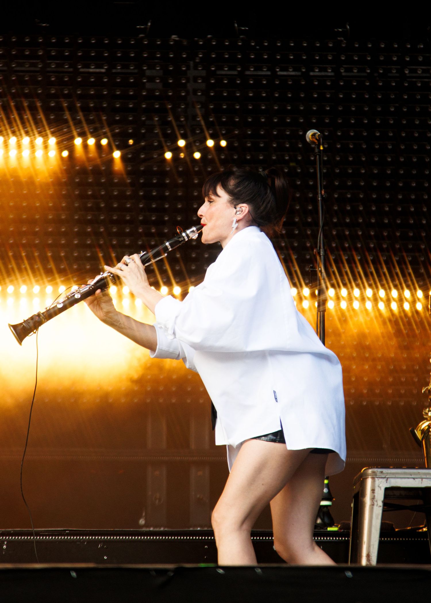 A performer in a white oversized shirt plays clarinet on a dramatic golden-lit concert stage.