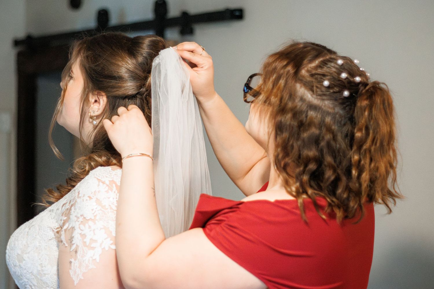Bridesmaid in red dress helping bride attach wedding veil to her hair before the ceremony.