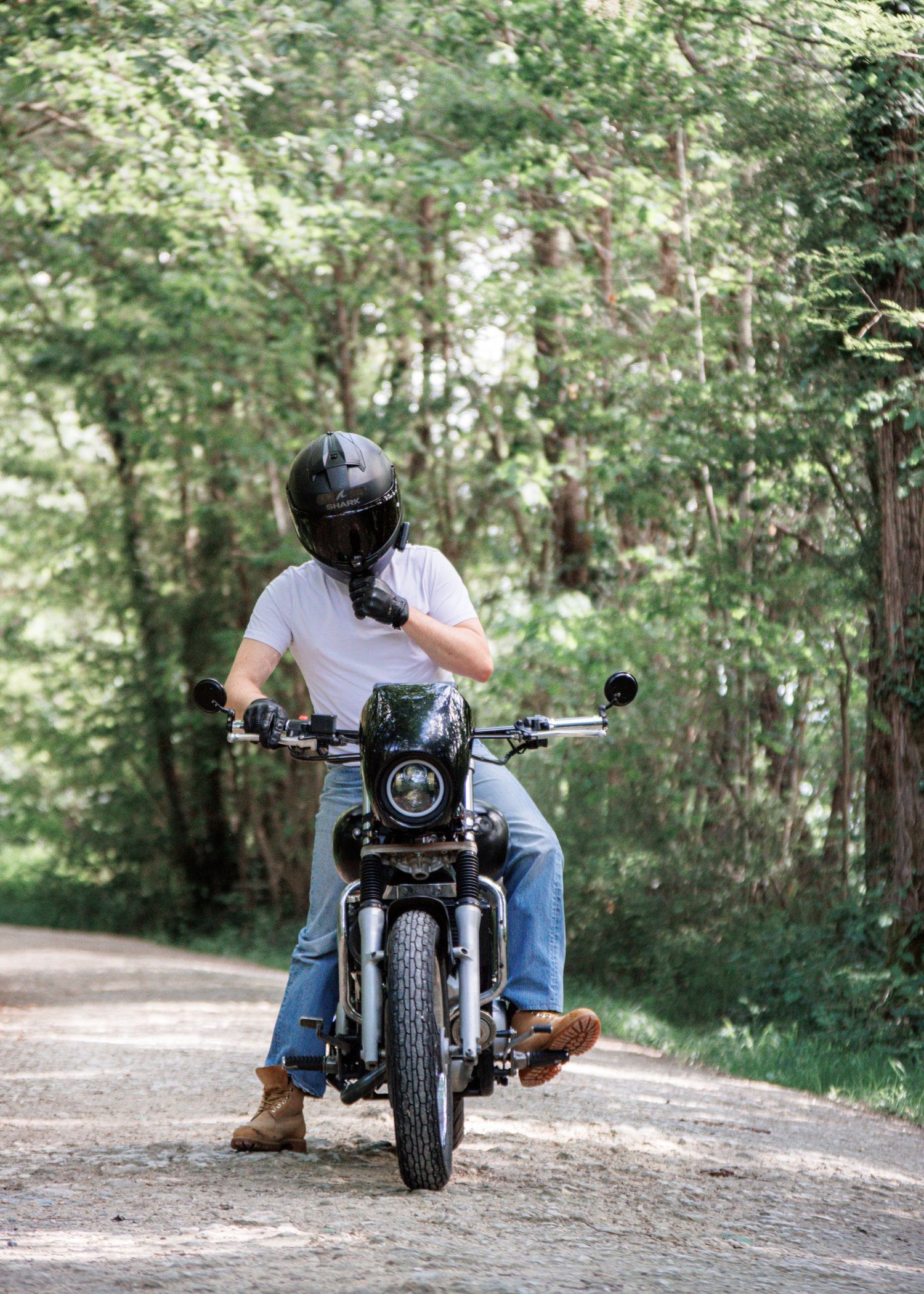 A motorcyclist in a white t-shirt, jeans, and black helmet sits on a motorcycle on a tree-lined road.