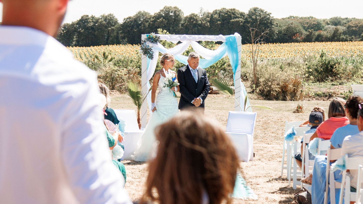 An outdoor wedding ceremony with white chairs set up in a field under a floral arch.