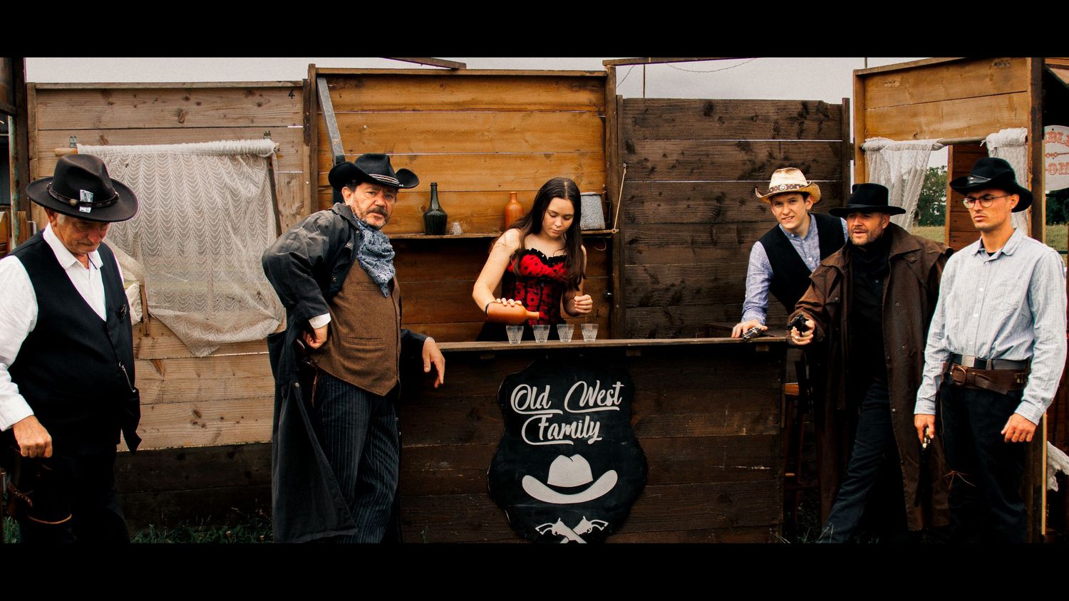 Group of people in western costumes gather around a rustic wooden bar setup.
