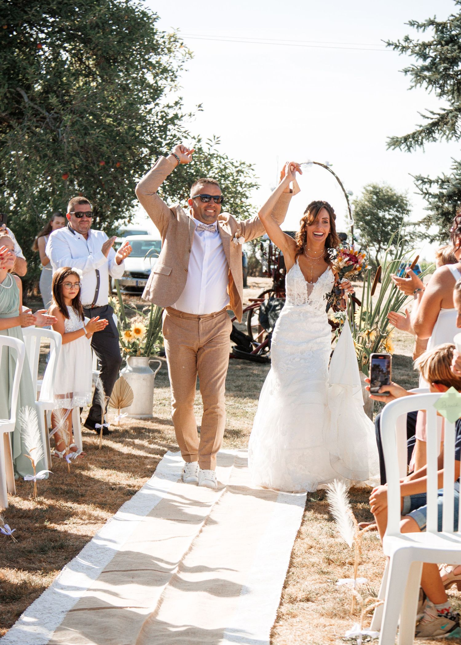 A couple celebrating at an outdoor wedding ceremony raise their hands triumphantly while walking down the aisle.