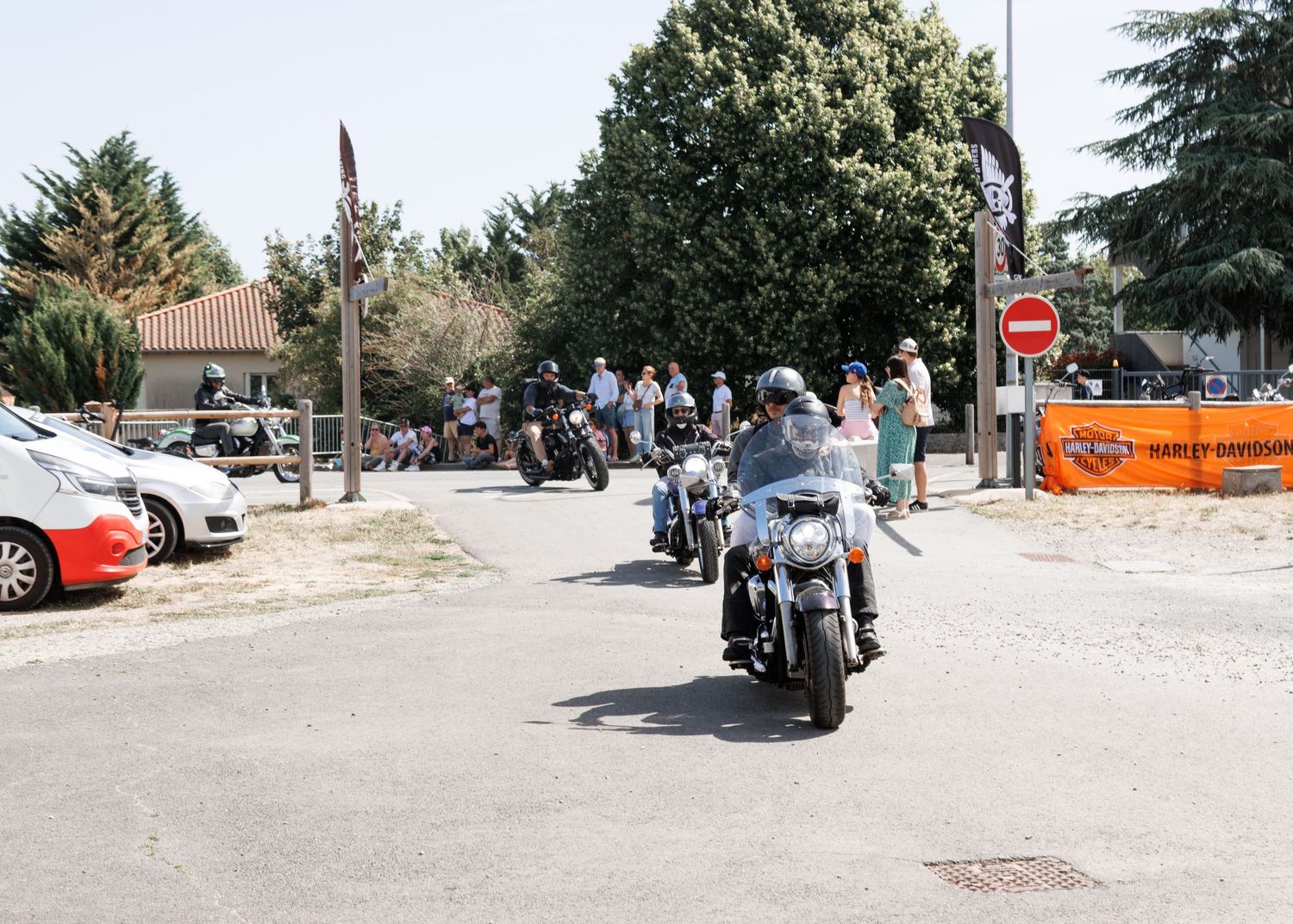 Motorcyclists riding past a Harley-Davidson banner on a sunny street with spectators and parked cars nearby.