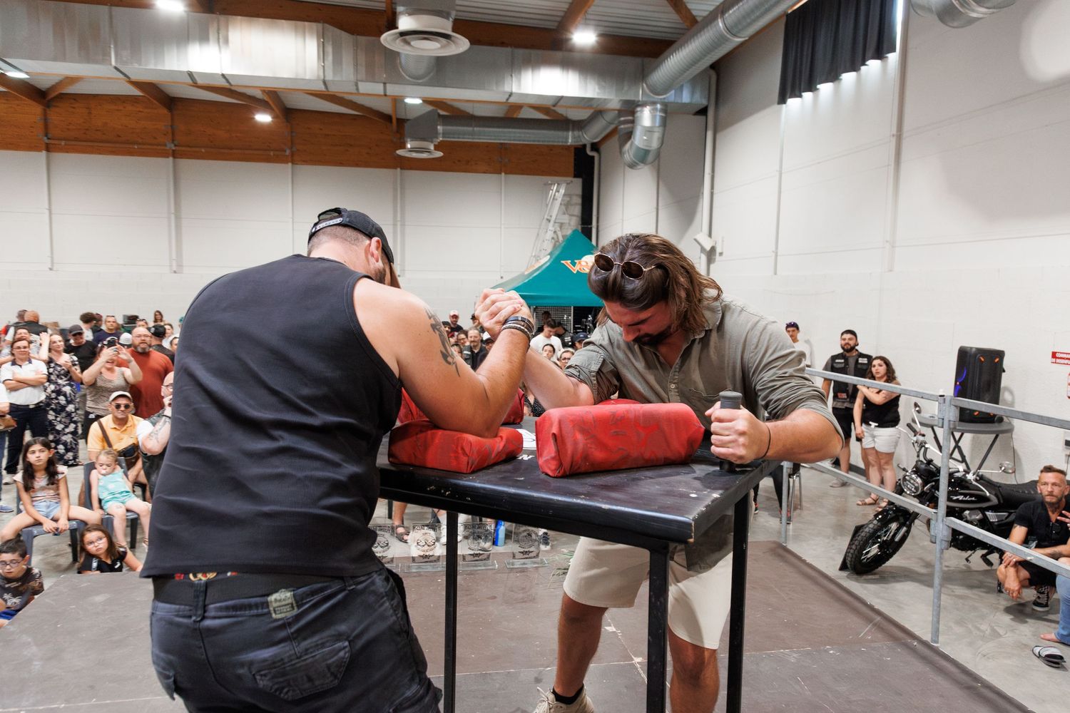 Two men competing in an arm wrestling match at an indoor event with spectators watching in the background.