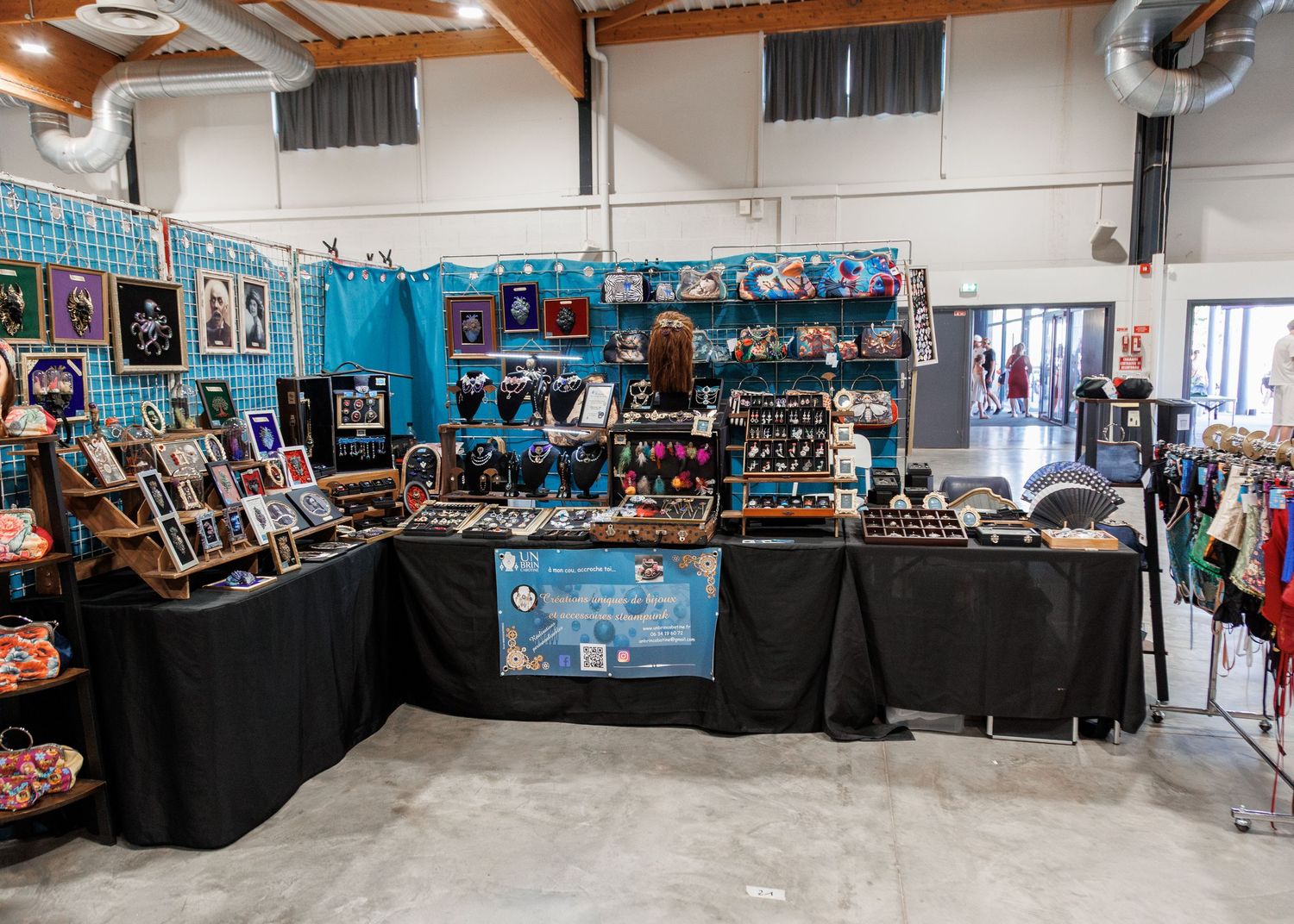 Vendor booth displaying handmade jewelry and accessories on black draped tables with teal backdrop at an indoor craft fair.