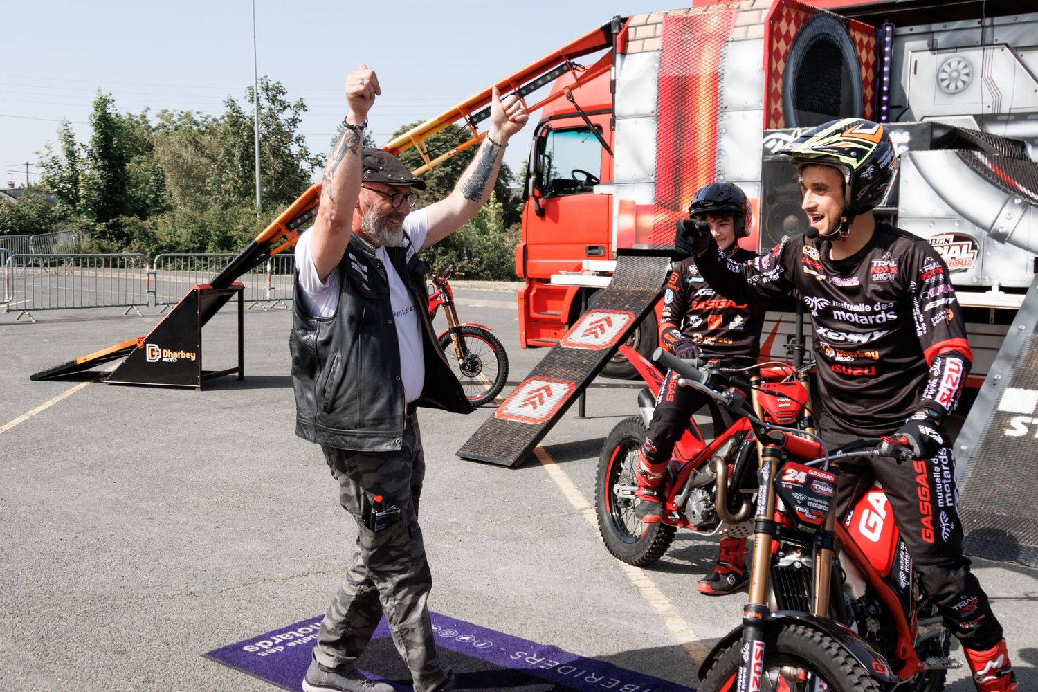 Two motorcycle riders celebrate with a team member raising his fist in victory at an outdoor motorsport event.