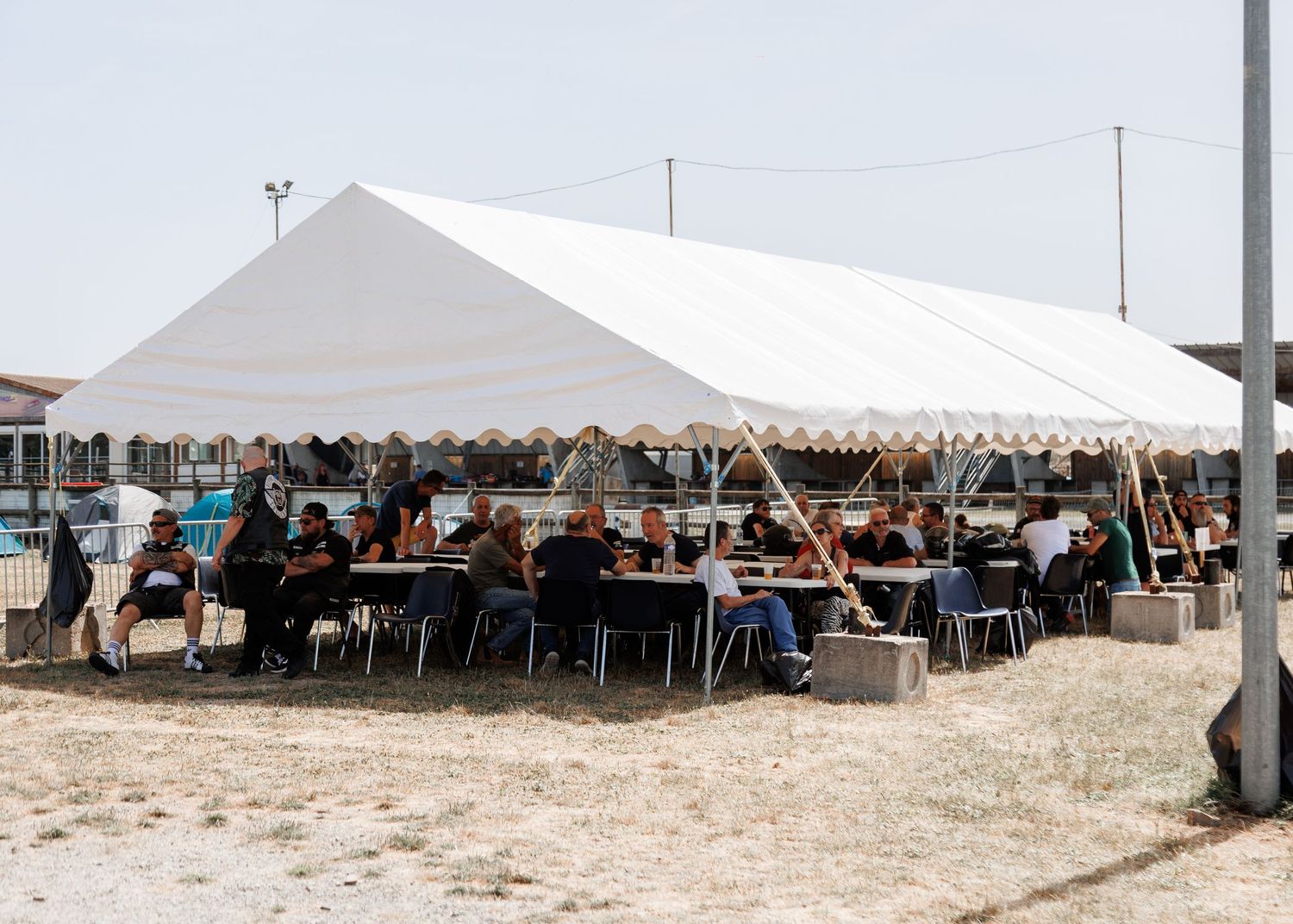People gathered under a large white outdoor tent with tables and chairs for an event on a sunny day.