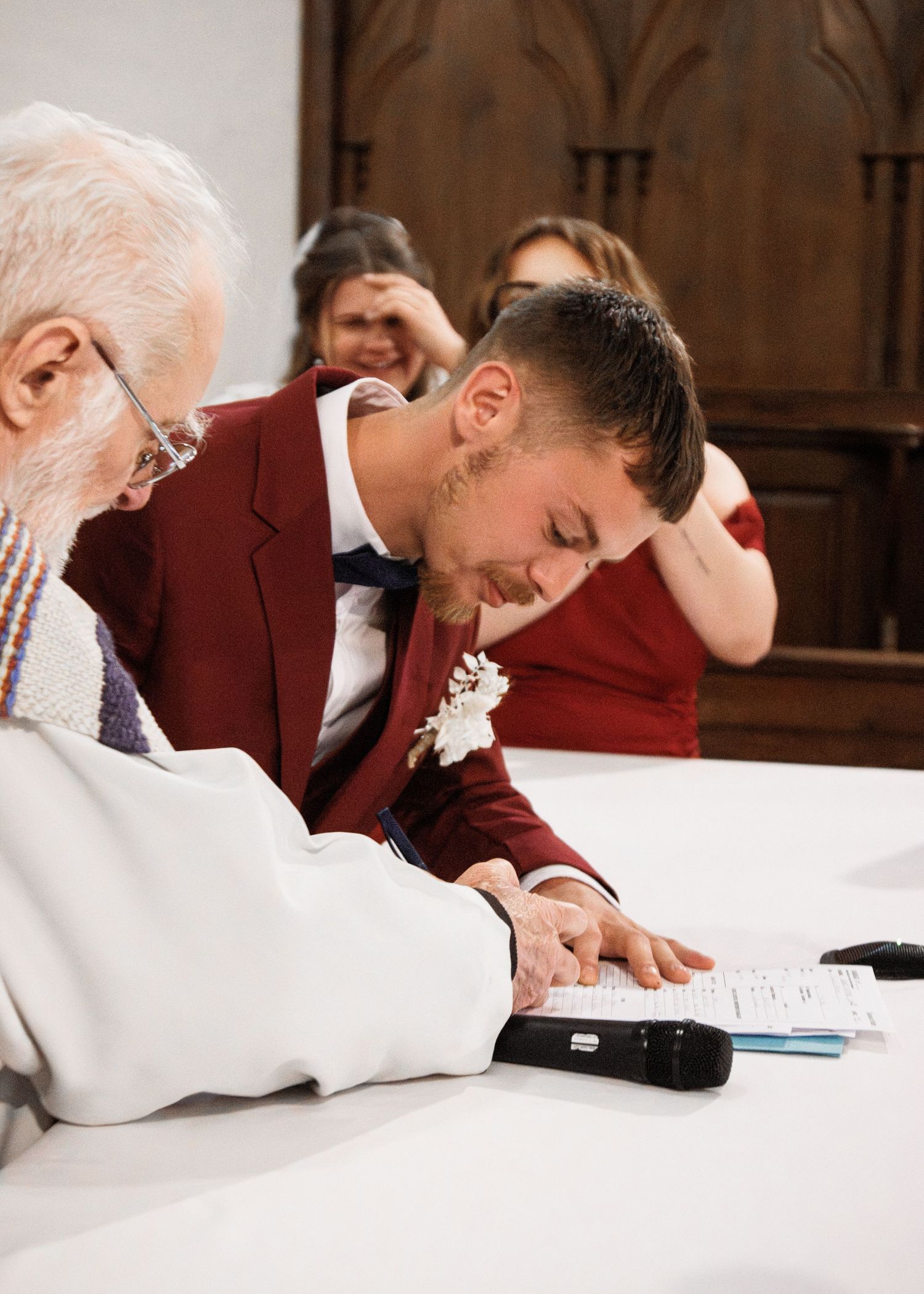 Groom in burgundy suit signing wedding documents at ceremony table with officiant.