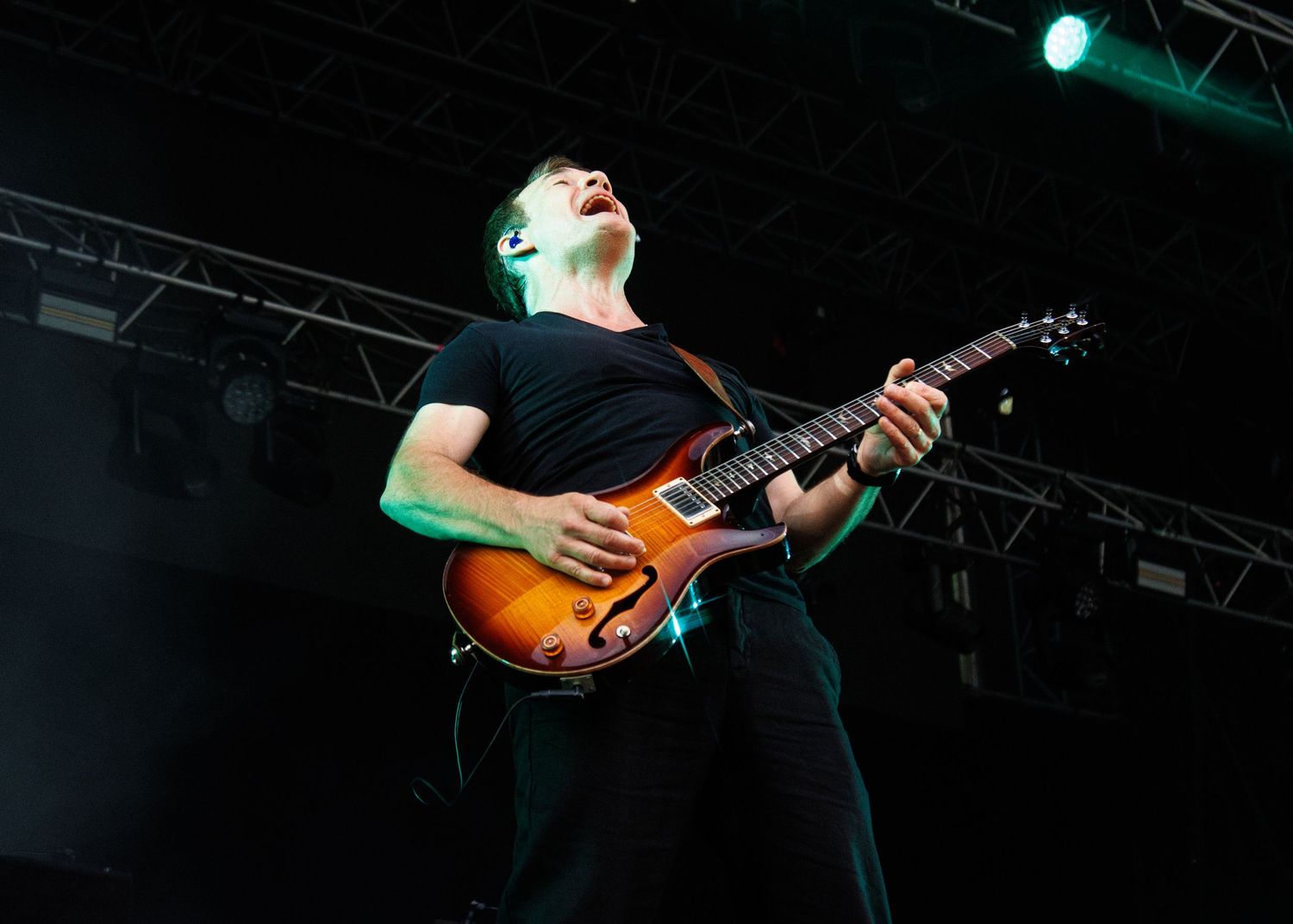 Guitarist performing on stage with electric guitar, wearing black outfit under concert lights.