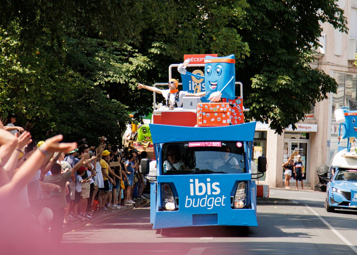 A blue Ibis Budget branded promotional truck drives through a crowded street parade.