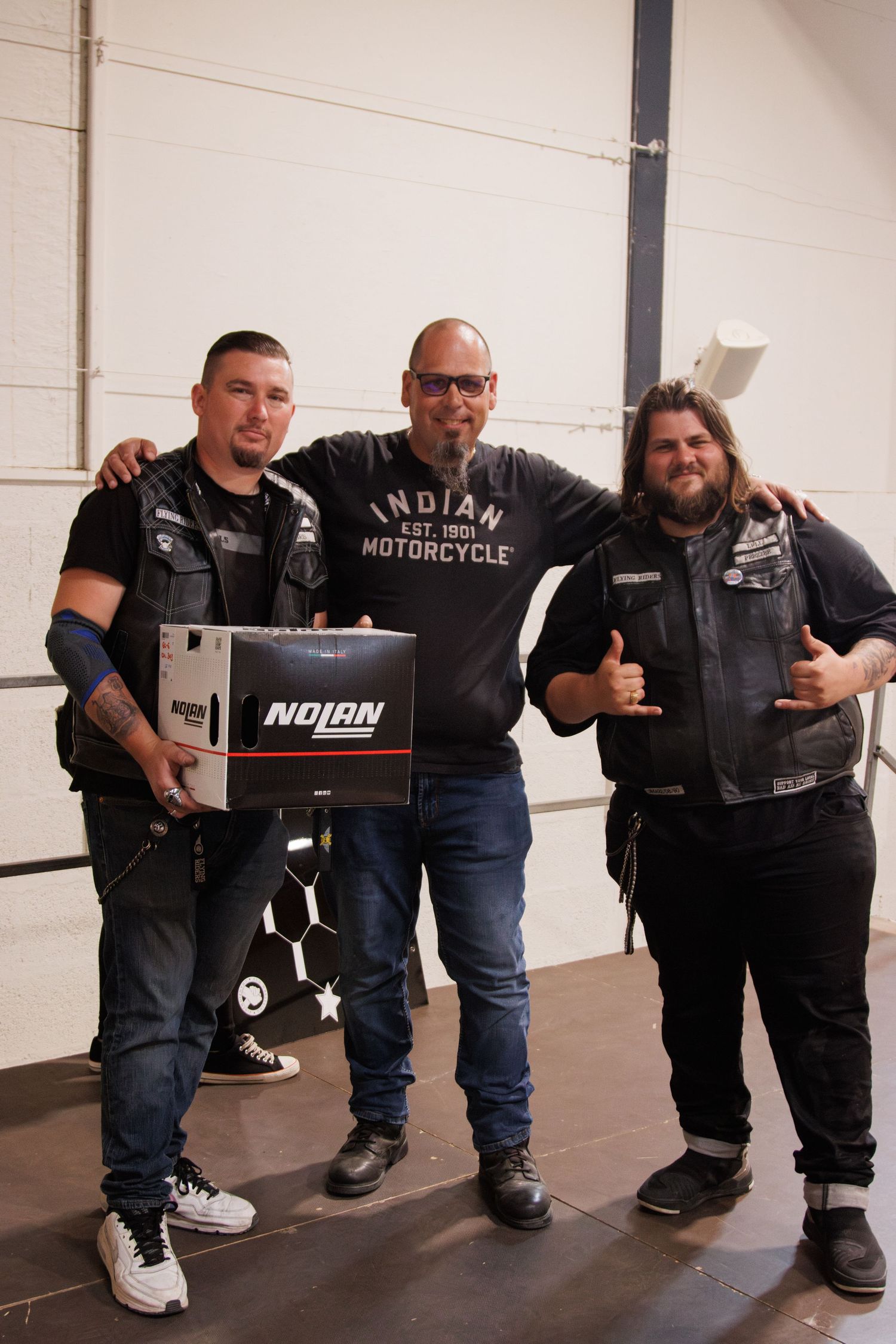 Three motorcycle enthusiasts stand together displaying Indian Motorcycle merchandise and giving thumbs up.